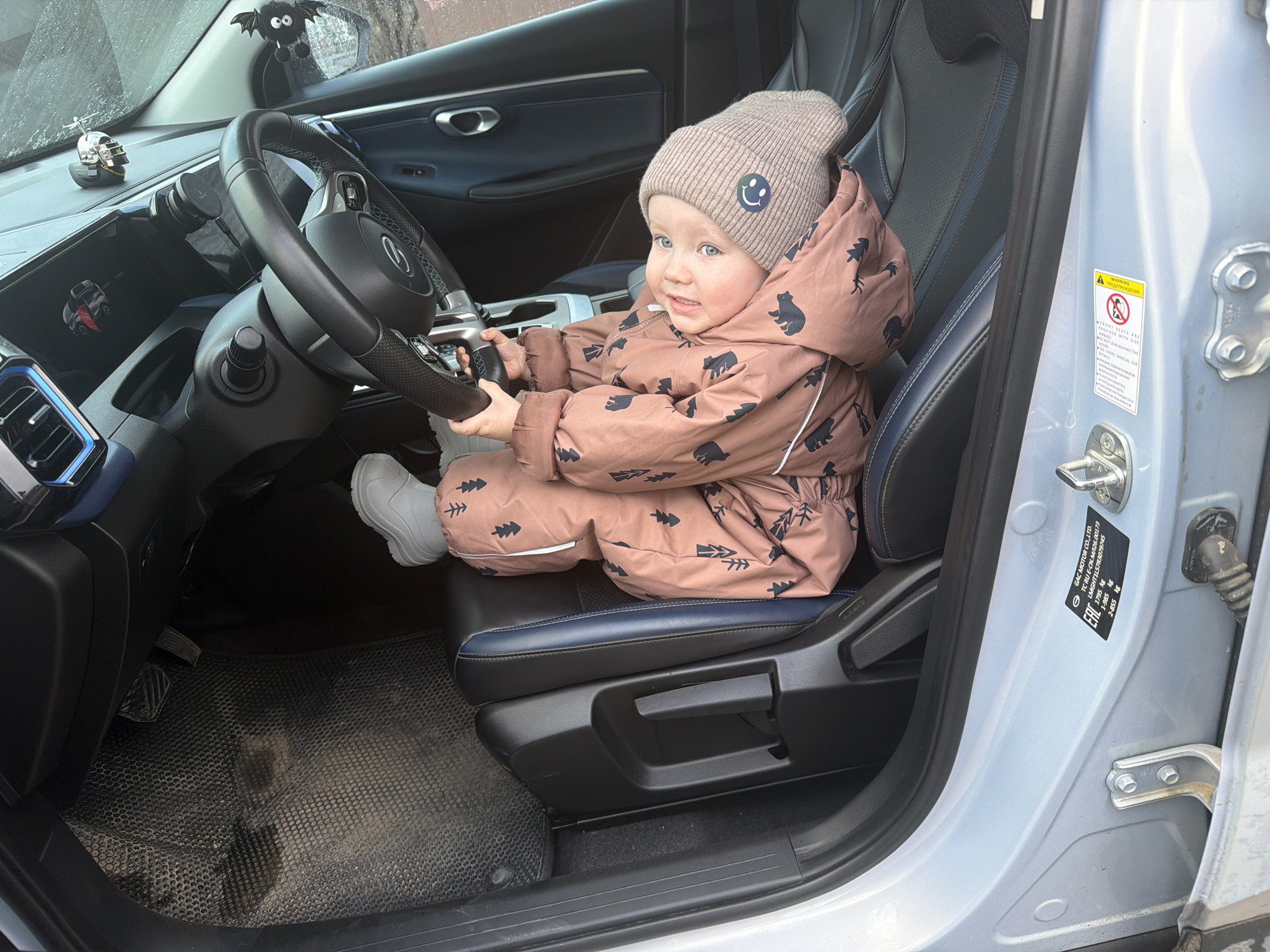 A young child, appearing to be a toddler, is playfully seated in the driver's seat of a car, gripping the steering wheel. The child wears a light brown knit beanie adorned with a blue smiley face and a brown winter jumpsuit featuring patterns of trees and bears, paired with light grey boots. They look towards the viewer's right with a curious and slightly smiling expression.
The car's interior includes dark leather seats with blue stitching and accents. The steering wheel prominently displays a Mercedes-Benz logo. The dashboard features a digital display, a large central screen, and a magnetic phone mount. Decorative accessories visible on the dashboard and windshield include a silver duck toy wearing a helmet and a black bat-shaped toy hanging from the rearview mirror. The black, textured floor mat on the driver's side appears used.
On the open car door frame, two informational labels are present. One is a warning label in English ("WARNING") and Russian ("ПРЕДУПРЕЖДЕНИЕ"), cautioning about front seats equipped with side airbags and advising against leaning on the door, with pictograms illustrating these warnings. The other label provides vehicle manufacturing details, stating "GAC MOTOR CO.,LTD.", "TC RU E-CN.M126.00173", "EAC", "LMGHT1L5783079745", and various weight specifications (1795 kg, 1-985 kg, 2-855 kg). The child's winter attire suggests a cold climate, aligning with the location context of Chaikovsky, Russia. The scene is illuminated by daylight.