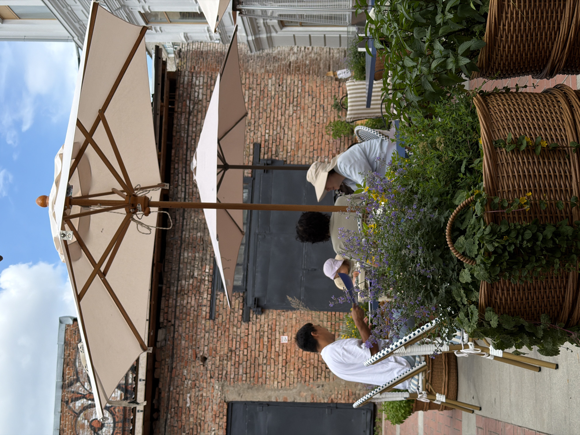 This image captures an outdoor scene with several individuals interacting amidst a charming setting. Two people are visible under a large, light-colored umbrella, one wearing a light-colored hat and the other a light blue shirt. Further to the right, another person in a white t-shirt is seated, holding a dark blue object, possibly a fan or a tool.
The scene appears to be a cafe or restaurant patio, with brick walls forming a rustic backdrop. Wicker baskets filled with lush green plants, including some with small purple flowers, add to the ambiance. The furniture includes chairs with black and white patterned detailing. The overall impression is one of a relaxed daytime gathering. The lighting suggests it's daytime, and the presence of plants and outdoor seating hints at pleasant weather. The specific location is Ufa, Russia. No visible text is present in the image.