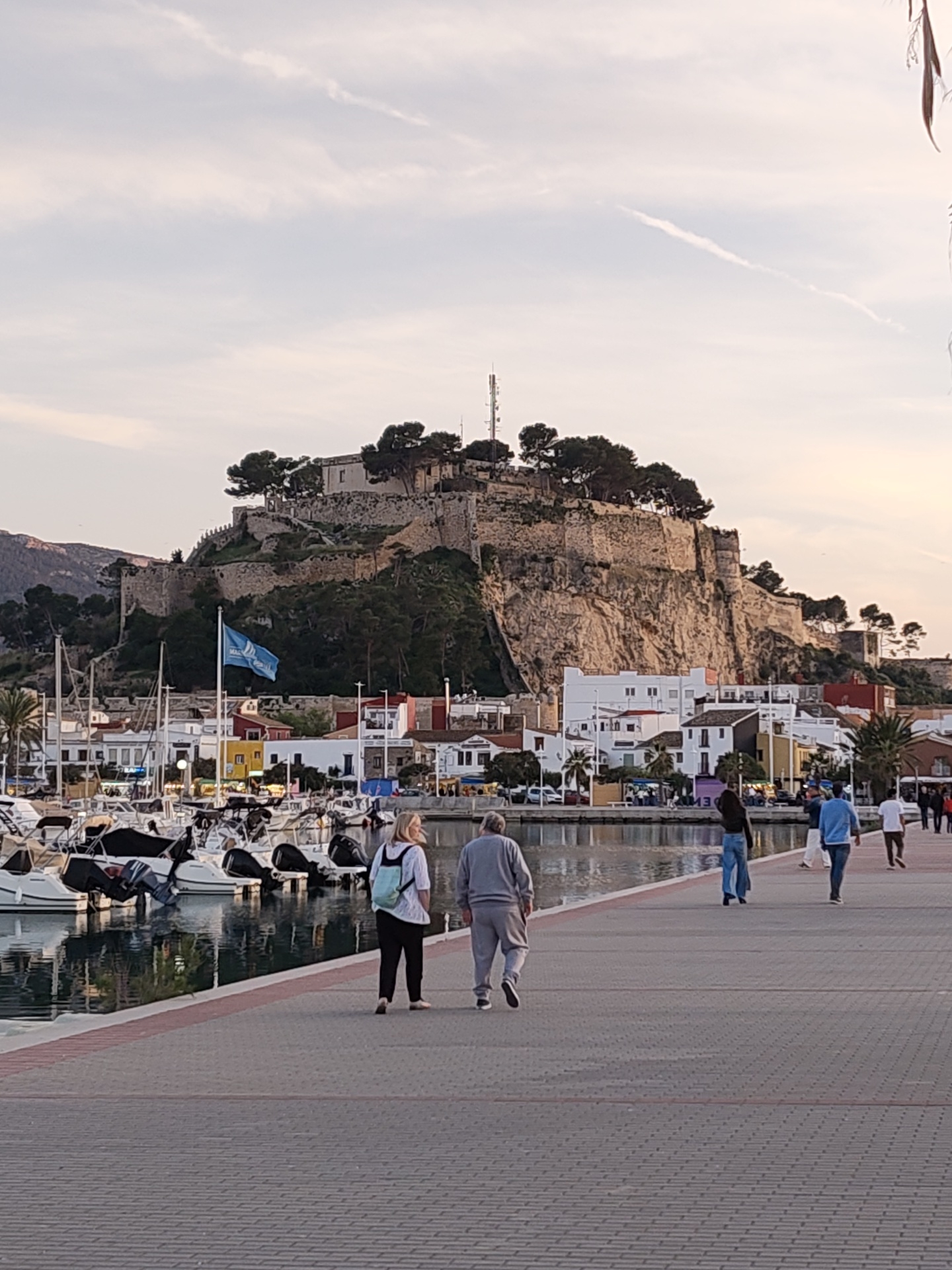 The image depicts a waterfront scene in Dénia, Spain, featuring a paved pedestrian walkway lined with boats docked in a harbor. In the background, a fortified structure, possibly a castle, sits atop a hill overlooking the town. Several people are walking along the walkway, including a couple in the foreground and other individuals further away. The buildings in the background appear to be a mix of residential and commercial structures. The sky is overcast with a visible contrail.