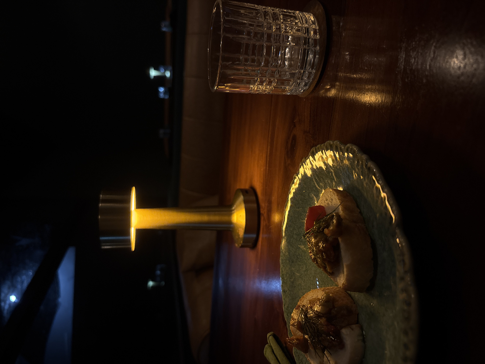 The image shows a bar scene in Kuta Selatan, Indonesia. On a dark wooden surface is a textured glass, a silver cylindrical lamp with a yellow light, and a light green plate with two appetizers. The appetizers appear to be topped with relish and herbs. The lighting is dim, with the lamp illuminating the plate and part of the wooden surface. The left side of the image is very dark. There are no people visible.