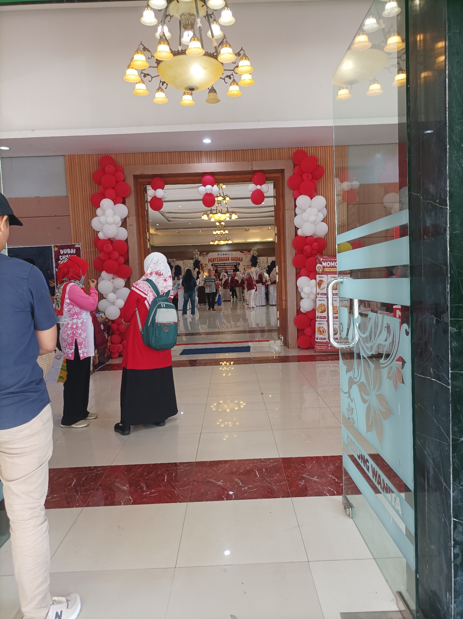 The image shows the entrance of a building decorated with red and white balloons. Inside, there are people, some wearing hijabs, gathered for an event. A banner that reads "Penyerahan Hadiah Festival Mewarnai" is visible, indicating a children's coloring festival award ceremony. The floor is tiled with a red and white pattern. A glass door with frosted floral designs and text "GEDUNG WANITA" (Women's Building) is on the right side of the image, suggesting the location is the "Gedung Wanita" in Nganjuk, Indonesia. There are chandeliers on the ceiling.