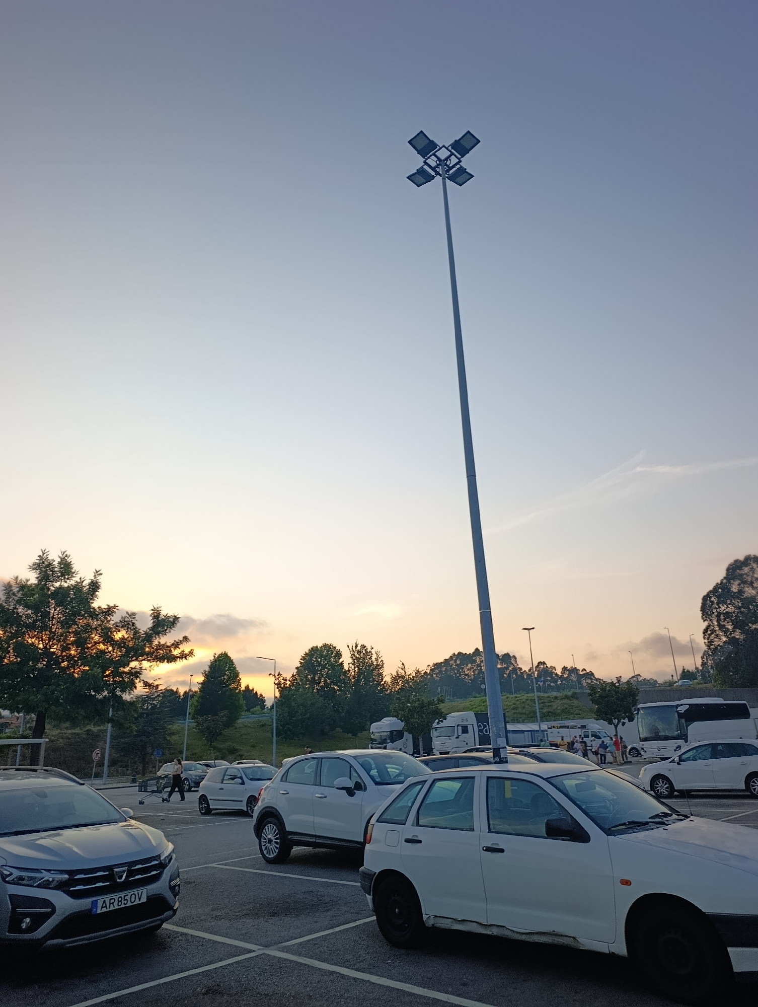A wide-angle outdoor, vertical, ground-level shot captures a parking lot in Santa Maria da Feira, Portugal, at dusk. The sky is a pale, hazy blue, transitioning to a warm orange and yellow near the horizon, indicating the setting sun. In the foreground, several cars are parked, including a silver Dacia with a Portuguese license plate "AR 850V" and a white hatchback. A tall, slender lamppost with four floodlights is prominently positioned near the center of the frame, extending high into the sky. Trees with dark green foliage border the parking lot, with some showing golden highlights from the sunset. In the background, more trees form a dense line along a gentle slope, and several other vehicles, including a white bus and trucks, are visible in the distance. The parking lot pavement is visible with painted parking lines. A few individuals are present in the scene, with one person pushing a shopping cart near a white car, and another person walking in the middle ground. The overall atmosphere suggests a public space, possibly a rest stop or commercial area, at the end of the day.