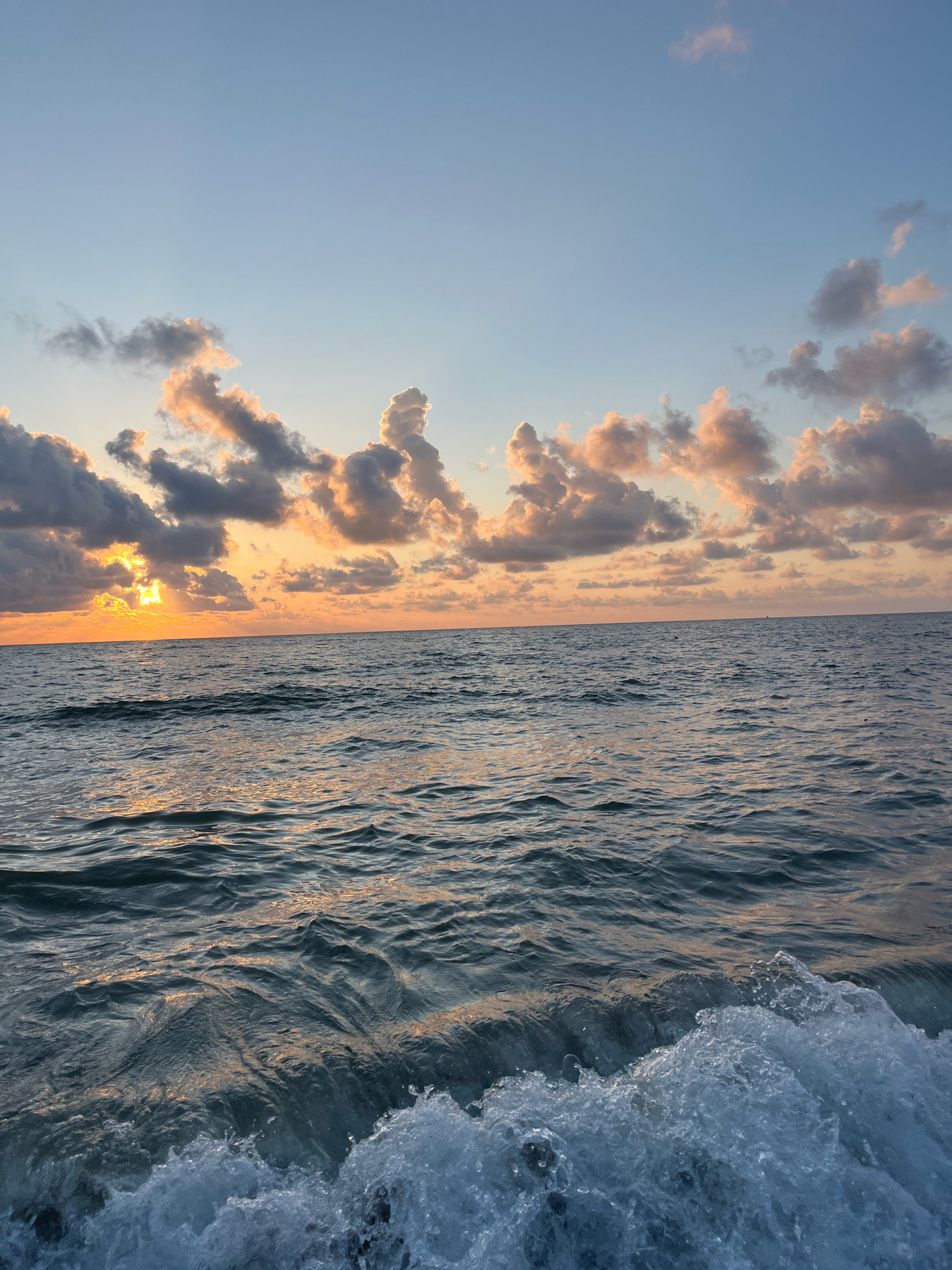 The image depicts a serene ocean scene at sunrise or sunset, likely in Ureki, Georgia, a town known for its Magnetic Sands beach. The sky is a blend of soft blues and warm oranges, with scattered clouds tinged with the golden hues of the rising or setting sun. The sun itself is partially obscured by clouds, casting a bright, radiant glow. The ocean surface is calm, with gentle waves creating ripples that reflect the colors of the sky. In the foreground, a wave is breaking, creating a frothy white crest of water. There are no people or specific man-made objects visible in the image, emphasizing the natural beauty of the coastal environment. The overall impression is one of tranquility and natural splendor.