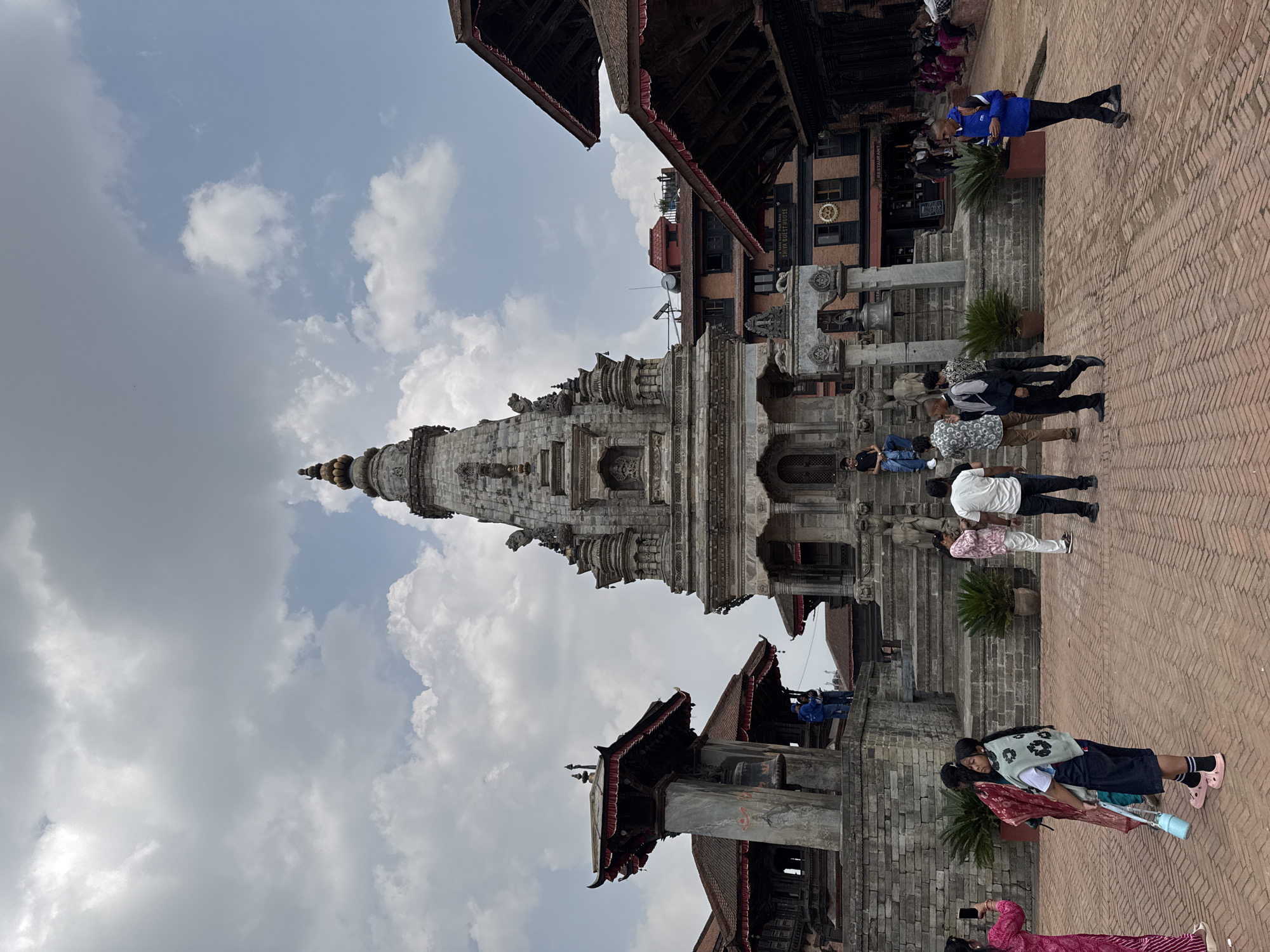 The image depicts a multi-tiered stone temple in Bhaktapur, Nepal, set against a cloudy sky. The temple is the main subject, and it features intricate carvings and a spire at its peak. Several people are present, some walking up steps towards the temple entrance, while others are standing or sitting nearby. The surrounding area appears to be a brick-paved public square with other traditional buildings visible in the background. A building with the sign "SIYA GUEST HOUSE RESTAURANT" can be seen. The overall scene suggests a tourist area with people visiting and exploring the historical site.