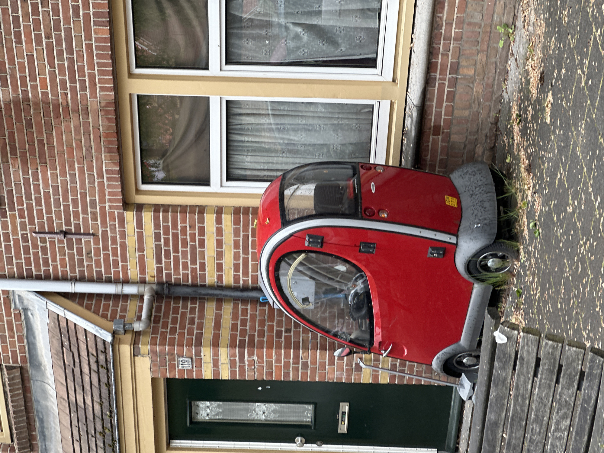 The scene is a street in Amsterdam, Netherlands, depicting a red mobility scooter parked partially on a wooden platform against the side of a brick building. The building features a green door with a rectangular window and a window above with white frames and drawn curtains. A gutter and pipe run along the brick wall. The ground is paved with patterned stones, scattered with leaves and small plants. The scooter is facing the right of the frame. The scene is captured in daylight.