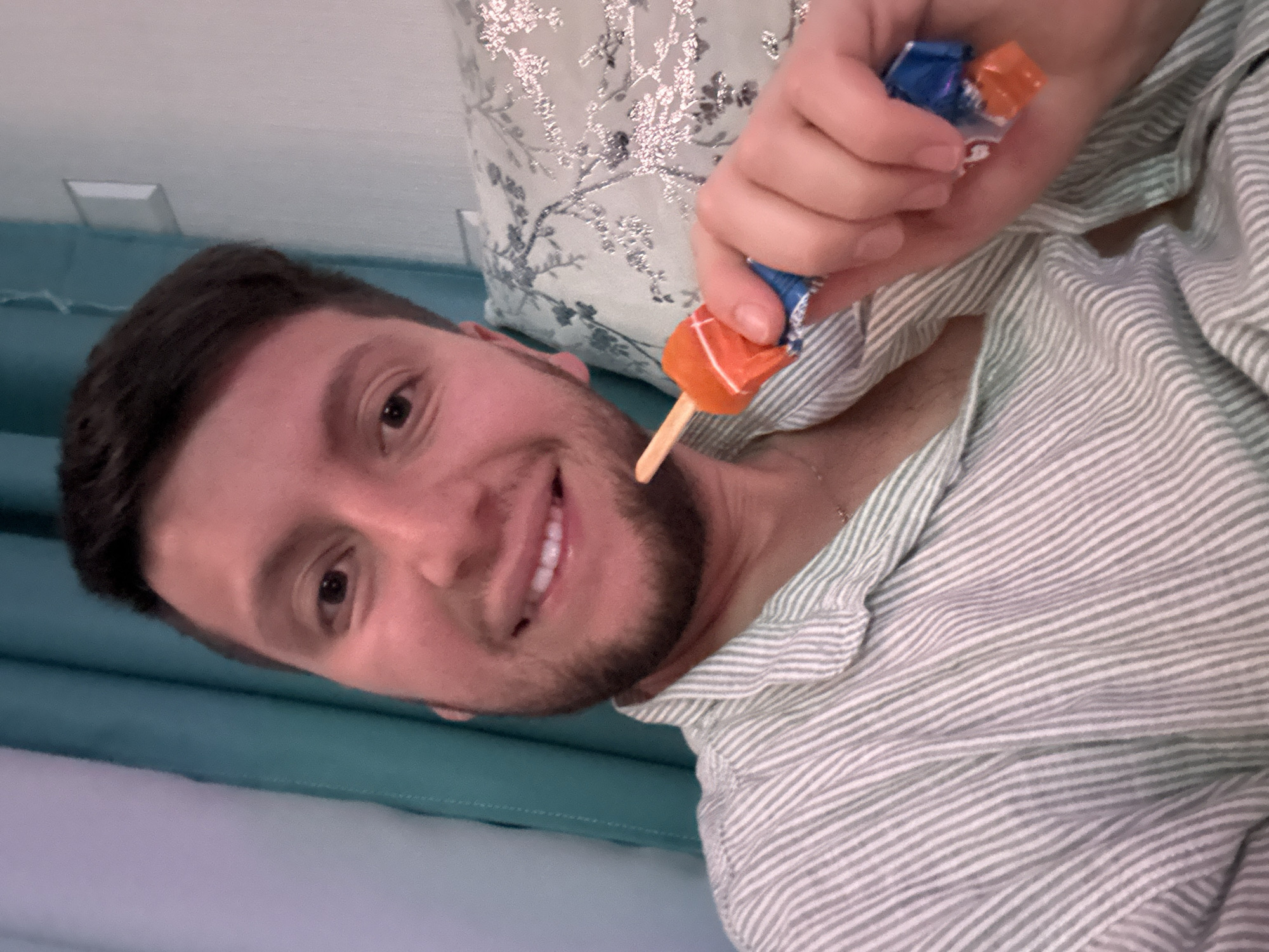 This image depicts a young adult male, of apparent Russian descent, approximately in his 20s or early 30s, located in the city of Мирный, Russia. He has short dark hair, light skin, and a neatly trimmed beard or stubble. He is smiling directly at the viewer, indicating a selfie or direct portrait. He wears a light-colored, possibly pale green or white, vertical striped button-up shirt. In his right hand, he holds a small confection on a wooden stick, partially unwrapped; the wrapper appears to be a combination of blue and orange. The background features a teal-colored curtain on the left side of the frame, alongside a lighter wall. On the right, a decorative pillow with a silver or grey floral pattern on a light background is visible, with a white wall and a light switch or outlet above it. The scene appears to be an indoor, relaxed setting.