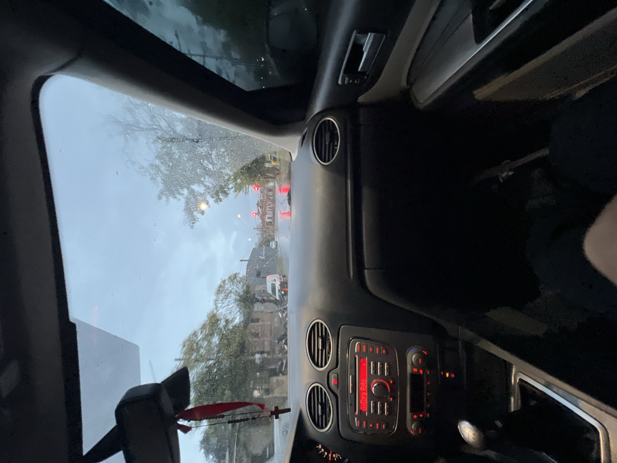 The image shows the interior of a vehicle, specifically the dashboard and windshield, taken from the driver's perspective. It is raining, as evidenced by the water droplets on the windshield and the overcast sky visible through the glass. The vehicle's dashboard features air vents and a car radio with a red display. Outside the windshield, there is a street scene with buildings, trees, and a white van, possibly in the city of San Fernando del Valle de Catamarca, Argentina. There are also traffic lights in the distance. A rosary is seen hanging from the rear view mirror. The focus is primarily on the scene outside the vehicle, framed by the interior elements. The overall environment appears to be urban or suburban, and the weather is a significant factor affecting visibility. There are no people visible outside the car. Inside the car, a dark leg and a part of a seat are slightly visible.