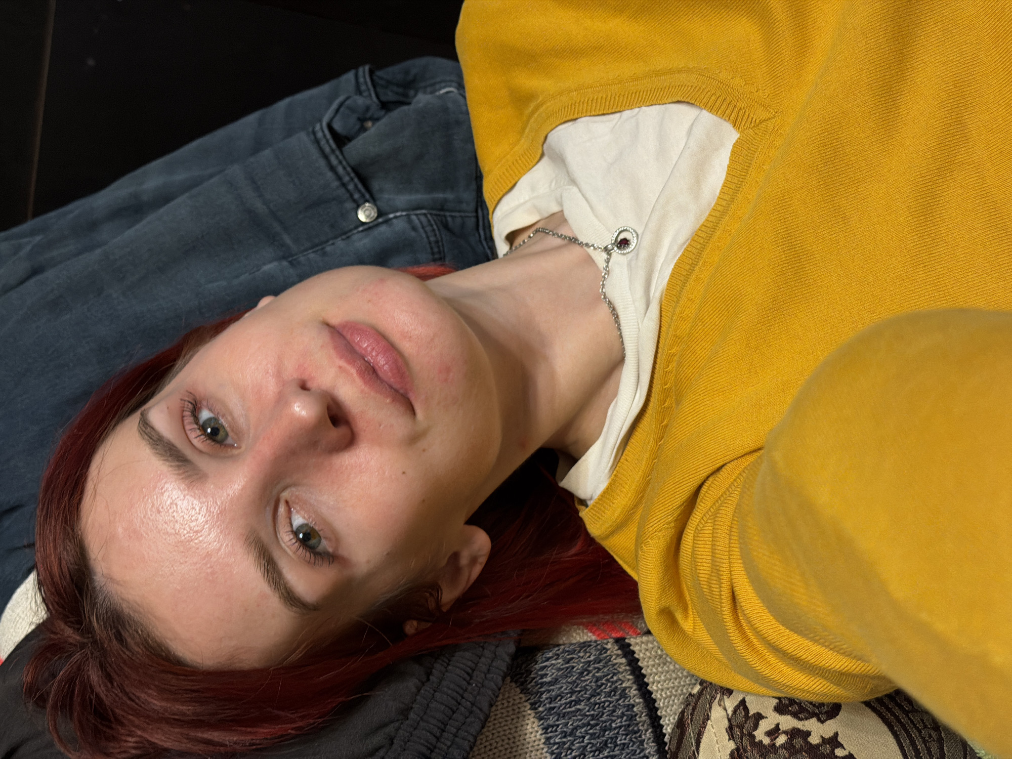 A close-up, rotated portrait captures a person, likely female, lying down indoors. The subject has reddish-brown hair and light-colored eyes, appearing to be green or hazel, looking towards the camera. Her complexion is natural, with minor redness visible around the chin and mouth. She is wearing a mustard yellow, ribbed long-sleeved sweater over a white undershirt. A delicate silver chain necklace with a small red or purple gemstone pendant rests on her chest.
To the upper left, a portion of blue denim jeans is visible. Below and to the right, a patterned fabric, possibly a blanket or cushion, suggests the person is resting on a soft surface. The lighting appears even, characteristic of an indoor setting. The subject's expression is relaxed and neutral. No text is visible in the image. The provided location context is Заболотово, Russia.