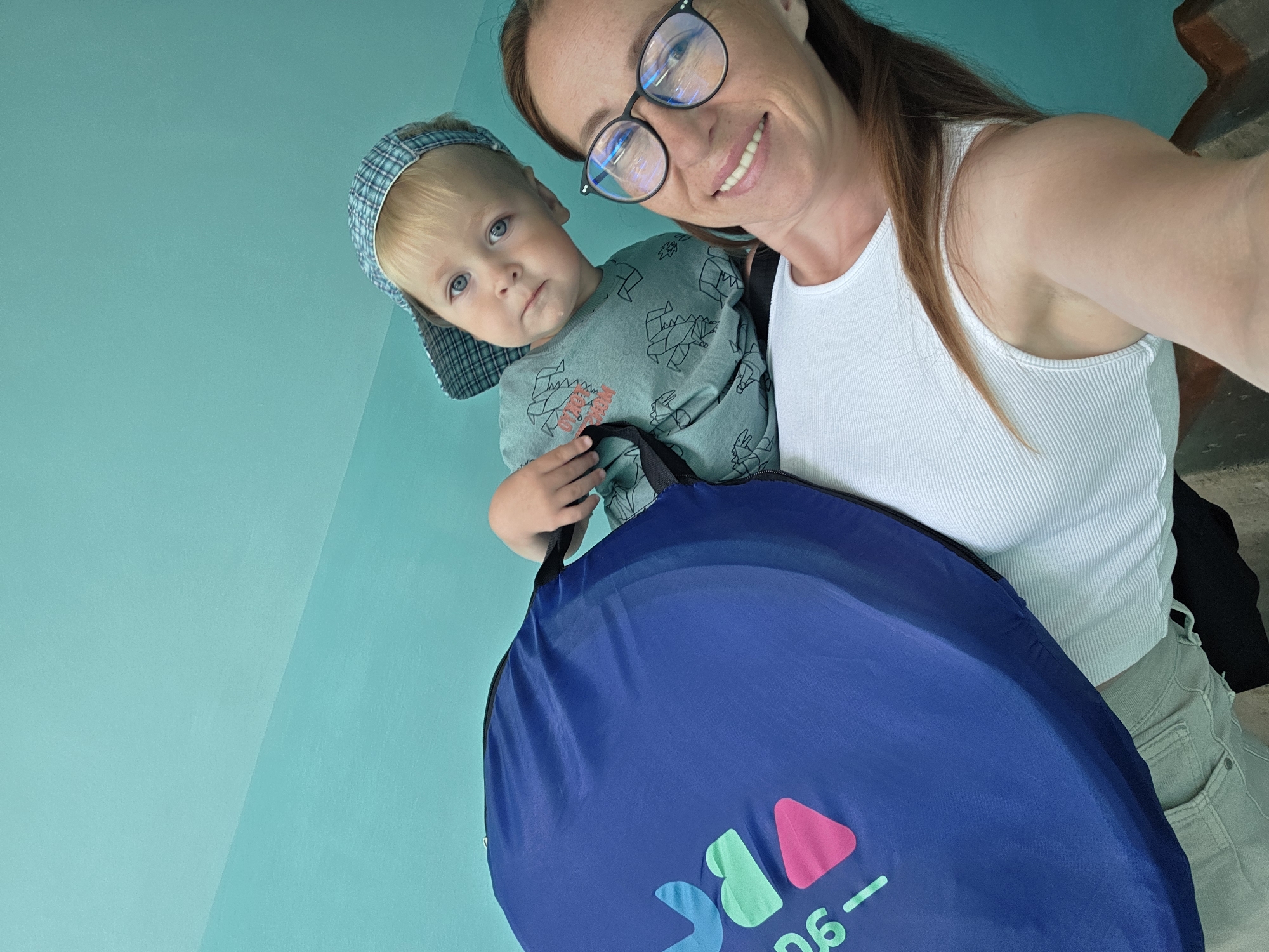 In a low-angle selfie taken indoors, a woman with long, reddish-brown hair and glasses smiles at the camera. She wears a white ribbed tank top and light-colored pants. A young boy with blond hair and blue eyes, wearing a blue plaid cap and a green t-shirt with a dinosaur print, is positioned behind her and slightly to her left. He is holding onto a large, dark blue circular bag with colorful graphics and text, including the numbers "96". The background is a solid, light blue wall. The overall impression is a candid, informal moment captured during domestic activity, possibly with a child's play tent. The location is not discernable from the image itself, and therefore cannot be confirmed to be in Chaikovsky, Russia.