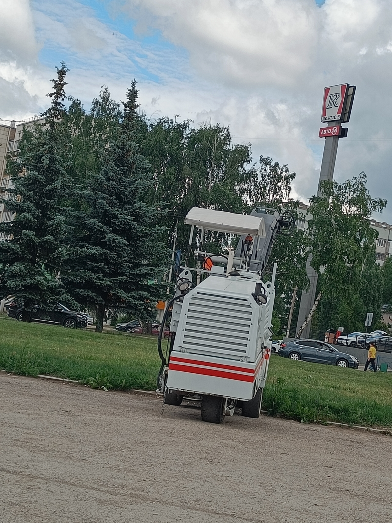 A white asphalt paver sits on a paved surface next to a grassy area. The paver is a large, industrial machine with a white body, red accents, and large wheels. In the background, several cars are parked on the side of the road, and a person in a yellow shirt walks along a sidewalk. A tall sign with a red "R" and "ROSTIC'S" written on it is visible behind the paver. Trees and apartment buildings can be seen in the background. The sky is cloudy with some patches of blue. The time of day appears to be daytime. The location appears to be Oktyabrsky, Russia.