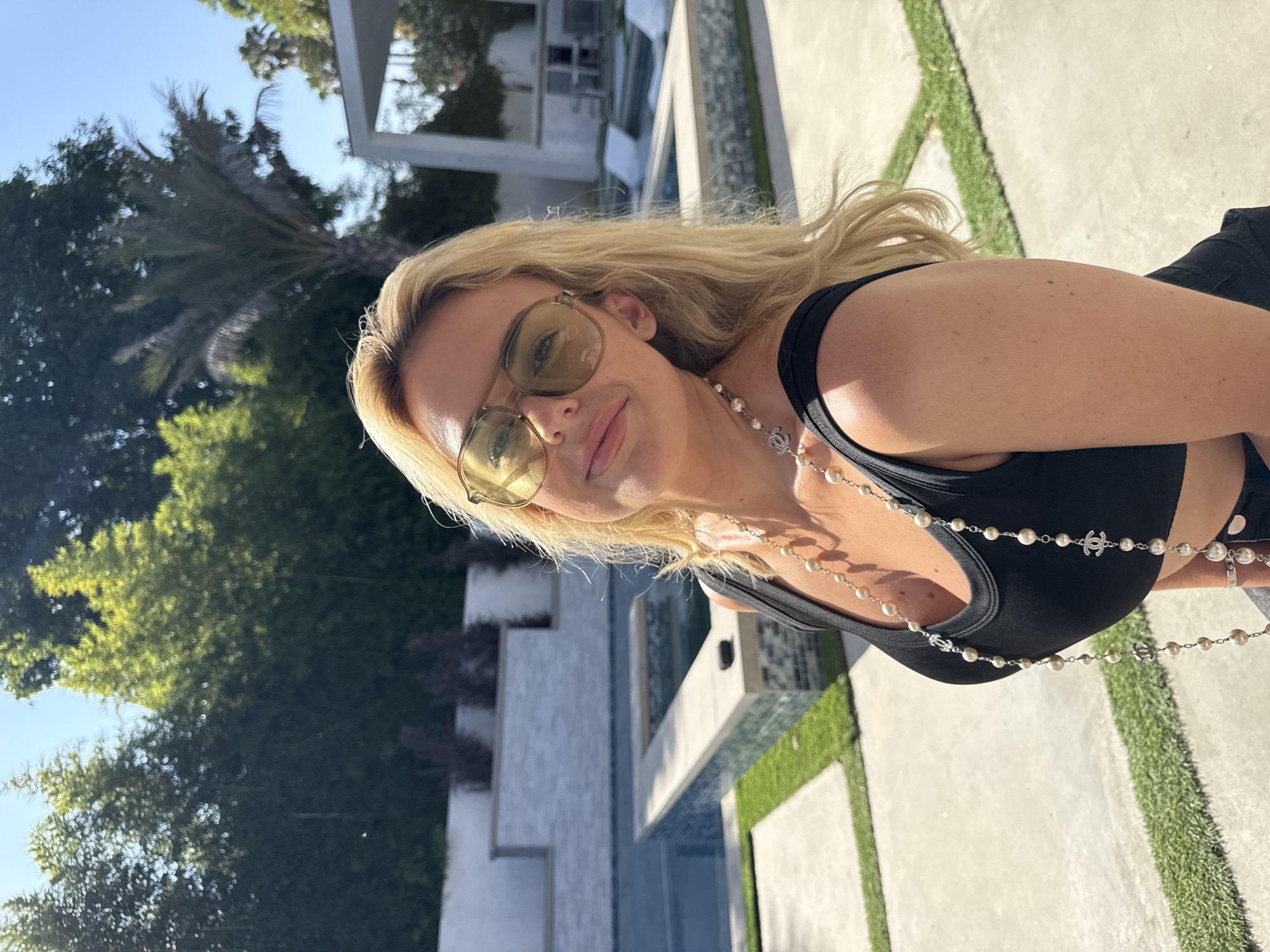 A woman with long blonde hair wearing yellow-tinted sunglasses, a black tank top, and a pearl necklace with Chanel logos is posing for a photo. She is outdoors on a sunny day, and the background shows a modern house with a pool and landscaping featuring grass, palm trees, and other greenery. The location is likely a residential area in Los Angeles, United States.