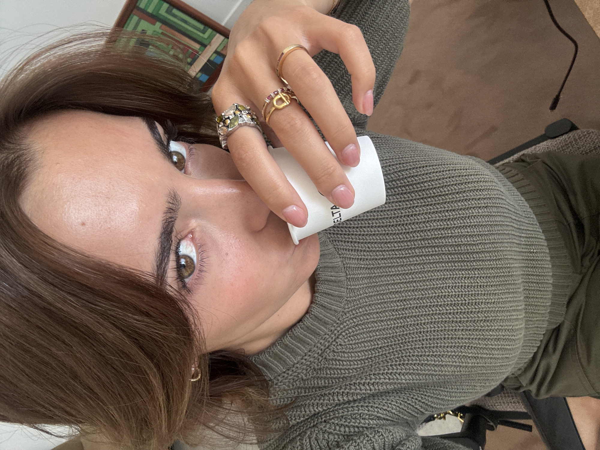 A woman with brown hair and light skin is holding a white cylindrical container with the word "DELTA" visible. She is wearing an olive green knitted sweater and several rings on her fingers. A small gold hoop earring is visible in her left ear. The background includes a partially visible framed artwork on the wall and a brown carpet. The specific location in Torres Vedras, Portugal, is indeterminable from the image.