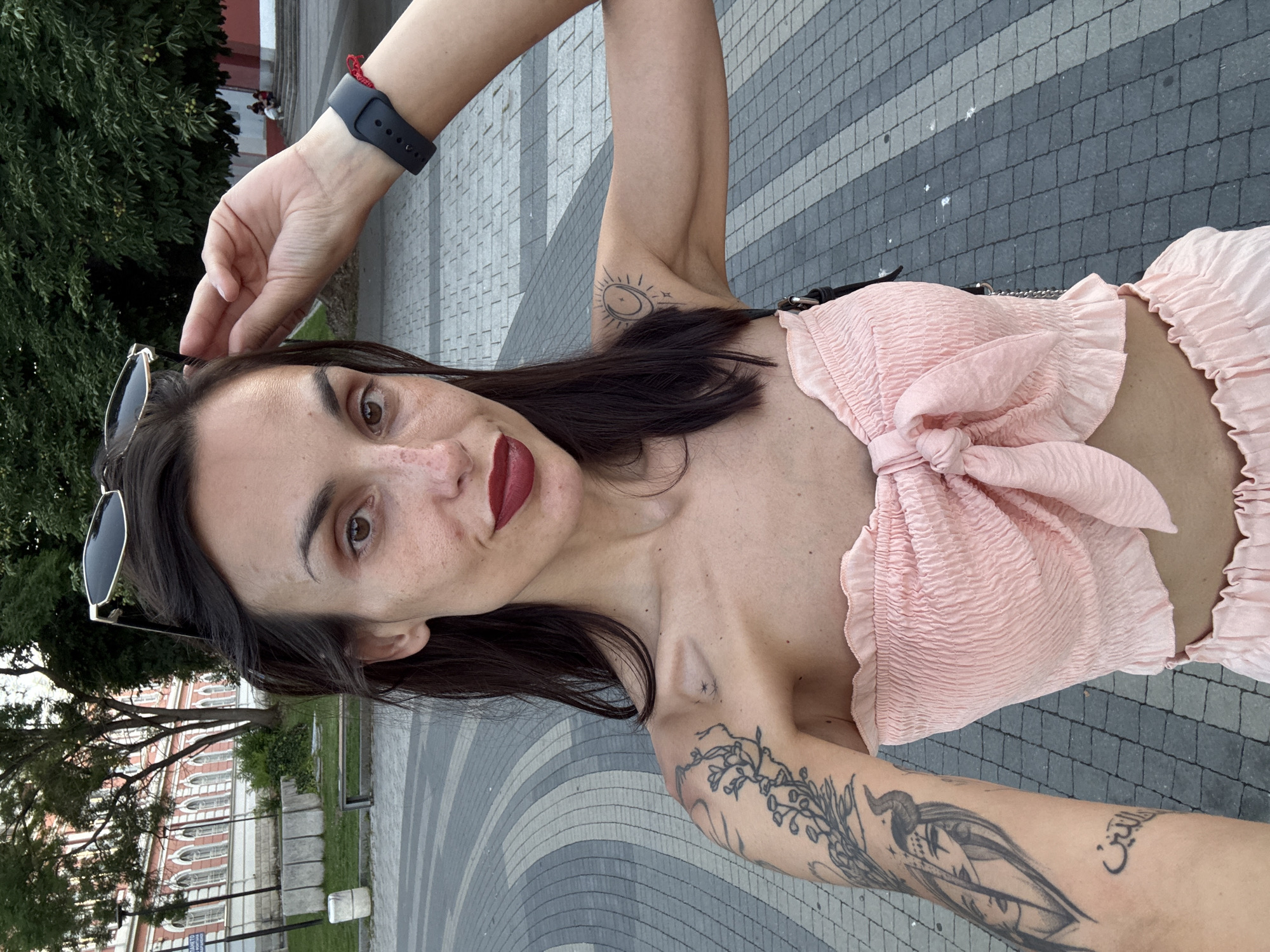 A woman is taking a selfie outdoors in Pleven, Bulgaria. She has long dark hair, freckles, and is wearing dark lipstick and a pink smocked tube top with a bow detail. She has tattoos on her left arm and shoulder, including a depiction of a face with horns and Arabic script. She is also wearing a black smartwatch with a red string bracelet. The background shows a paved area with a patterned design, a building with red and pink accents, trees, and a grassy area with a bench and concrete bollards. The woman's left arm is raised, touching her head, while her right arm is slightly bent. The image appears to be taken in the late afternoon or early evening, as indicated by the lighting.