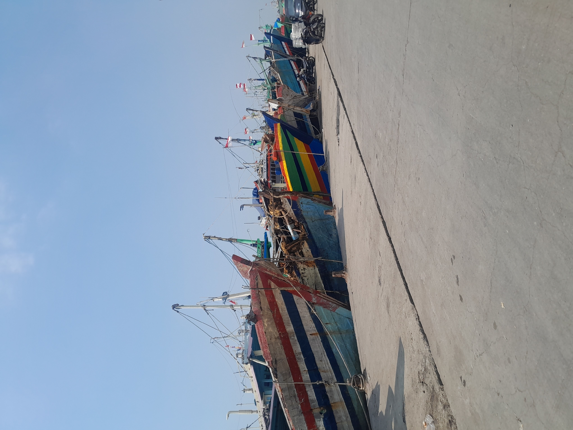 The image depicts a line of colorful fishing boats docked along a concrete pier in Tegal, Indonesia. The boats, varying in size and color, are tied to mooring posts. Some boats are painted with bold stripes of red, white, and blue, while others display vibrant rainbow patterns. The sky above is a clear, pale blue with a hint of white clouds. On the pier, several motorcycles are parked, and a black car is visible in the distance. The overall scene suggests a busy fishing port under a bright, sunny day.