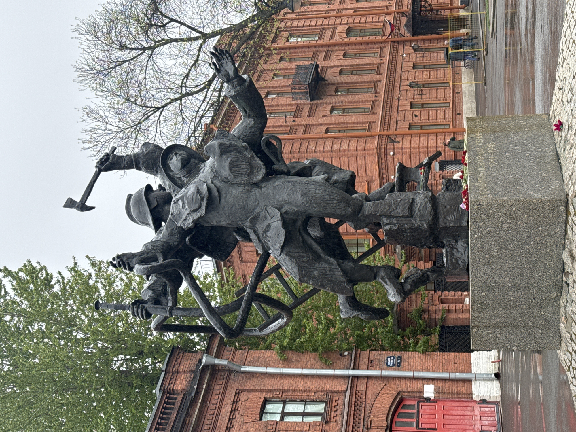 The image depicts a bronze monument in Saint Petersburg, Russia, in front of a red brick building with windows and a small balcony. The monument features three figures of firefighters with tools, including a ladder, axe, and hose. The firefighters appear to be in action. The monument rests on a stone base and is surrounded by trees and greenery. The sky is overcast, and the ground appears wet.