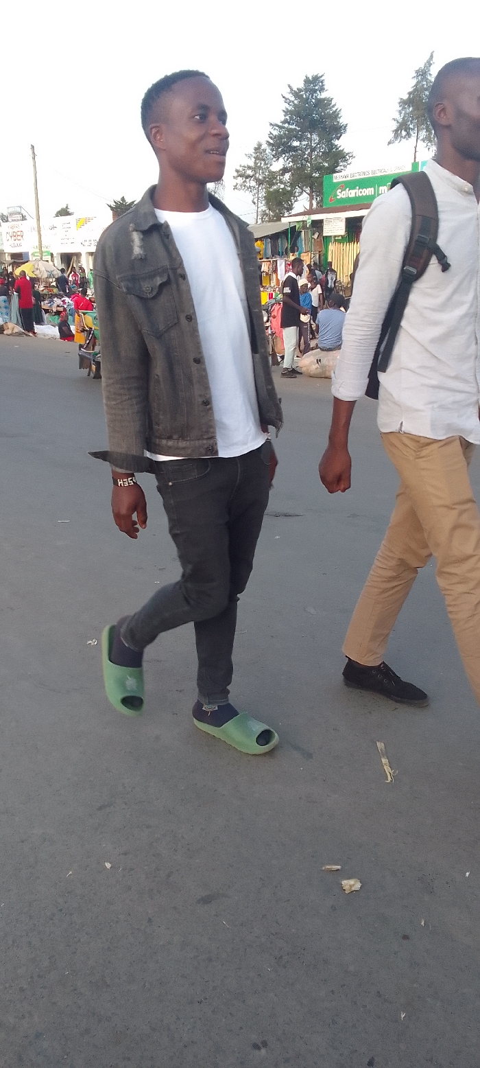 The image captures a street scene in Kakunga, Kenya, likely in a bustling market or commercial area during late afternoon or early evening. The soft lighting suggests this time of day. The weather appears clear and bright.
In the foreground, two men are walking on a paved road. The man on the left, closer to the camera, is young with dark skin and short dark hair. He wears a dark grey distressed denim jacket over a white t-shirt, dark jeans, dark blue socks, and light green slide sandals. A dark wristband with the white text "HSE" is visible on his left wrist. He is smiling or smirking, looking towards his right, suggesting he is engaging with someone or something off-camera. A dark object, possibly the handle of a knife or tool, is tucked into the left side of his waistband.
Partially visible on the right is another man, also with dark skin, wearing a white long-sleeved shirt, khaki trousers, and black shoes. He has a dark backpack and is walking in the same general direction as the first man.
The background is a lively market setting with numerous people, market stalls, and temporary structures lining the street. Various goods, possibly clothing and other items, are displayed in the stalls. Visible signs include "Safaricom m..." (referring to Safaricom, a major Kenyan telecommunications company) and "MEBAYA ELECTRONICS ELECTRICALS & HARDW...". Another partially obscured sign mentions "CYBER...". The scene depicts everyday activity in an African urban or semi-urban environment.
