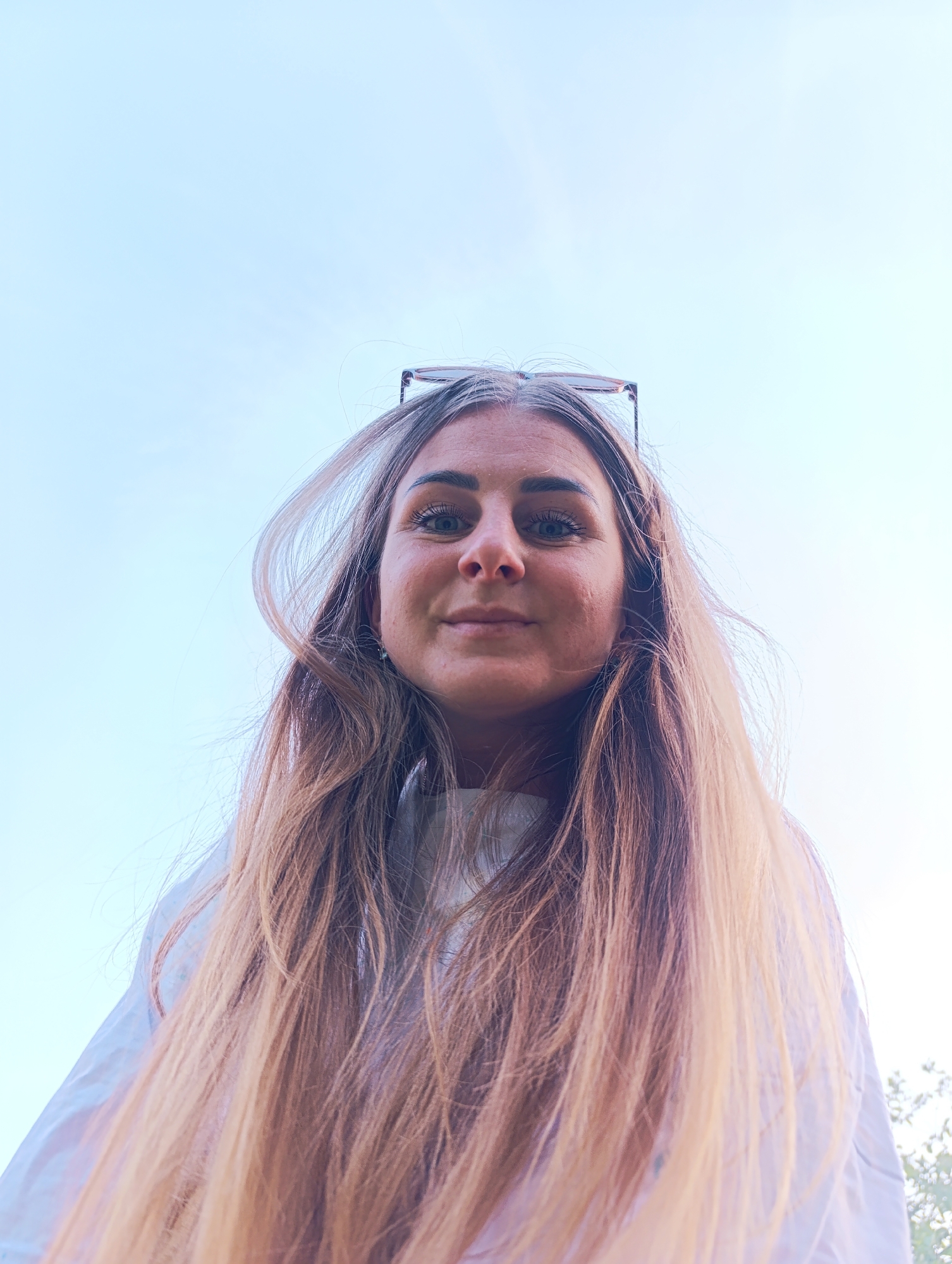 A woman is centered in the frame, looking down at the camera. Her long, light brown hair frames her face. She has a fair complexion and her eyes are blue. Glasses are perched on top of her head. She is wearing a white shirt. The background is a pale blue sky, suggesting an outdoor setting. The location is unknown.