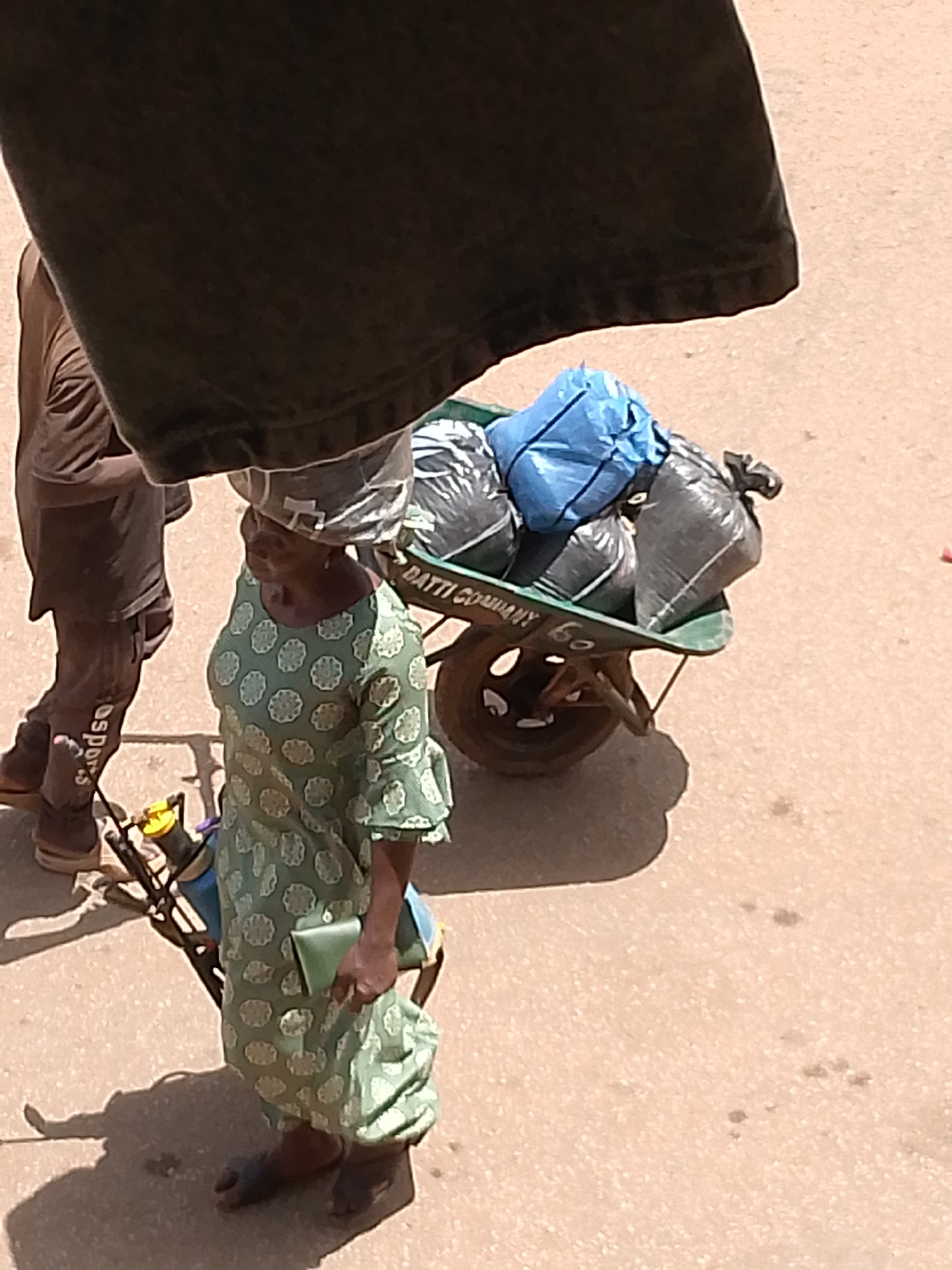 The image, apparently taken in Bukuru, Nigeria, presents an elevated view of a street scene. A woman is seen in the foreground, pushing a green wheelbarrow. The wheelbarrow contains black and blue bags. The woman is wearing a green patterned dress, a head wrap, and sandals. She is holding a green wallet or clutch in her hand. Next to the woman is a person who has brown sportswear pants. A dark object, possibly a piece of clothing, obscures the top portion of the image. The ground is paved and appears light brown or beige in color.