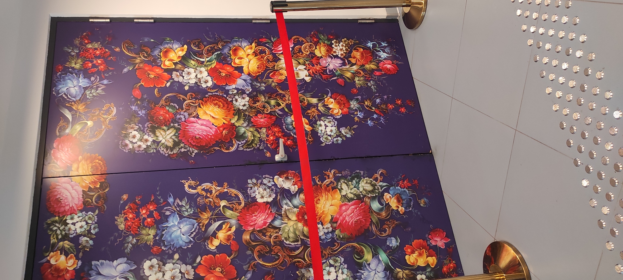 The image shows two tall, closed doors decorated with a vibrant, detailed floral pattern on a dark purple background. The pattern features large, rich-colored flowers such as peonies, roses, and other blossoms, interspersed with smaller flowers, leaves, and ornate gold scrollwork. A bright red ribbon is stretched horizontally across the lower portion of the doors, acting as a barrier. To the right of the doors, a stanchion with a gold pole and base is visible, also holding a red ribbon. The floor is tiled in a light gray color, and in the foreground, a line of raised tactile paving is present. The image appears to be taken indoors, possibly in a public space or entryway, and no people are visible. The specific location within Moscow, Russia cannot be determined from the visual information provided.