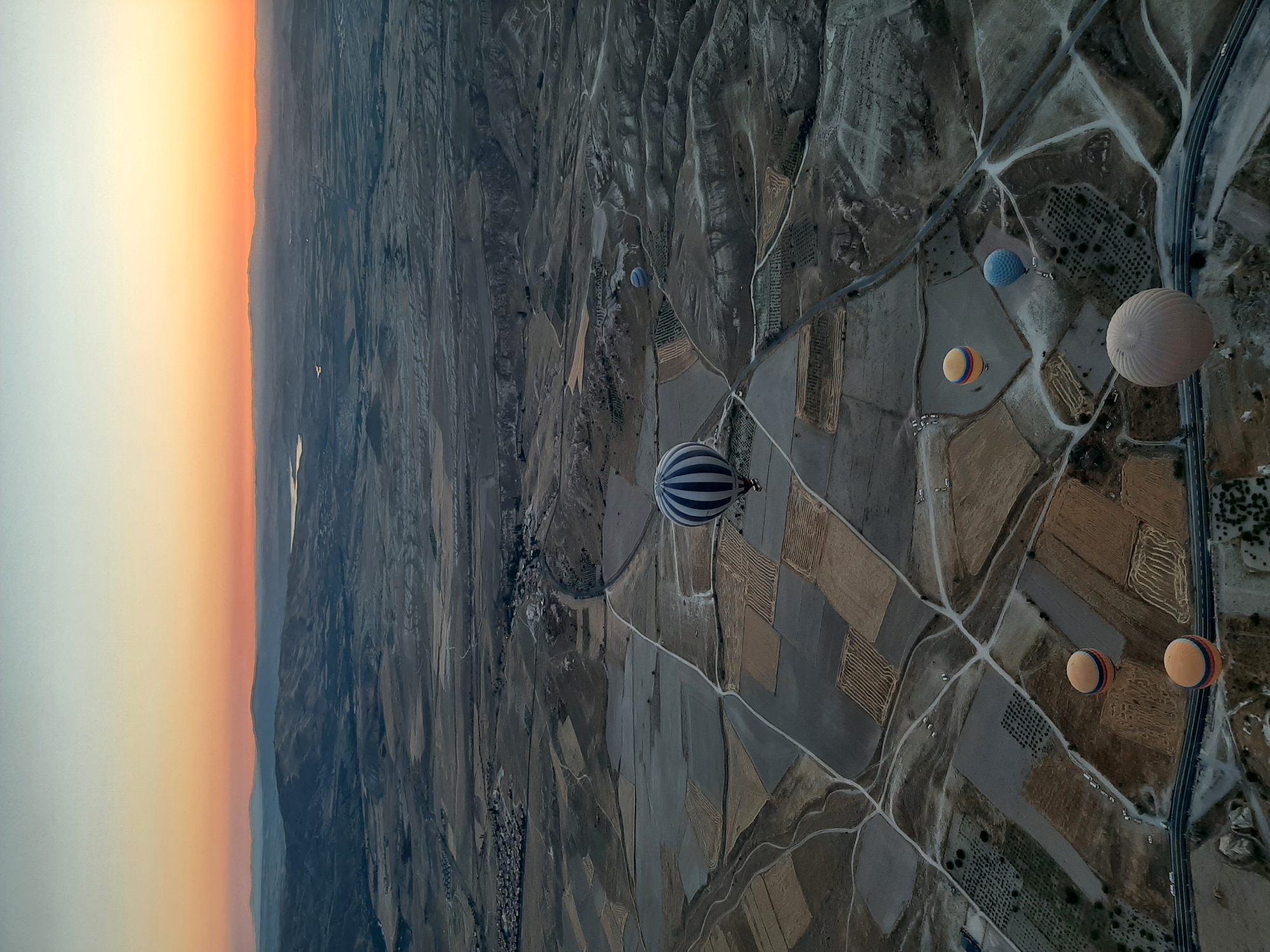 This aerial photograph captures the dawn sky over Avanos, Turkey, with hot air balloons dotting the landscape. The sky transitions from a pale blue at the top to a vibrant orange and pink near the horizon, illuminating rolling hills and valleys. The terrain below is a patchwork of agricultural fields, some fallow and others cultivated, divided by dirt roads and pathways. A winding highway runs through the lower portion of the frame. Several hot air balloons, varying in size and color, are visible: one large balloon with blue and white stripes dominates the center-left of the image, while others in shades of yellow, blue, and beige are dispersed across the scene. The balloons are floating at different altitudes, some appearing quite close to the ground and others higher in the sky. The overall impression is one of a serene and picturesque sunrise over a rural landscape.