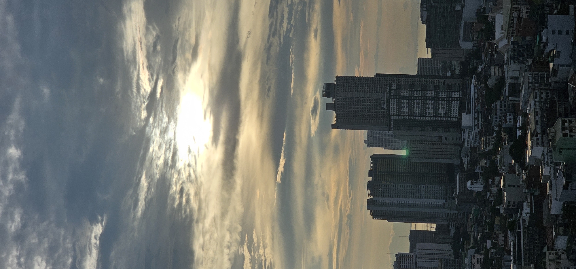 The image depicts a cityscape with a dramatic sky. In the foreground, a dense urban environment is visible, characterized by numerous buildings of varying heights and architectural styles. Many of these structures appear to be residential or commercial, with a mix of modern high-rises and smaller, older buildings. Some of the taller buildings exhibit visible construction or unfinished sections.
The middle ground is dominated by several prominent skyscrapers, some of which are quite tall and modern in design. One particularly large building in the center appears to be under construction, with visible scaffolding and an unfinished top. A distinct green light flare is visible on one of the buildings in the middle ground, possibly from camera lens or a reflection.
The background features a hazy horizon, suggesting the presence of water or a distant landmass, possibly the Manila Bay area, which is consistent with the location of Makati. The sky is filled with a multitude of clouds, ranging from dark, heavy masses to thinner, wispy formations. The sun is partially obscured by these clouds, creating a diffused, golden light that illuminates parts of the sky and casts a warm glow over the scene. The overall impression is one of a bustling city at either dawn or dusk, with the dynamic sky adding a sense of atmosphere and grandeur. The scene is devoid of any discernible people or specific actions taking place.