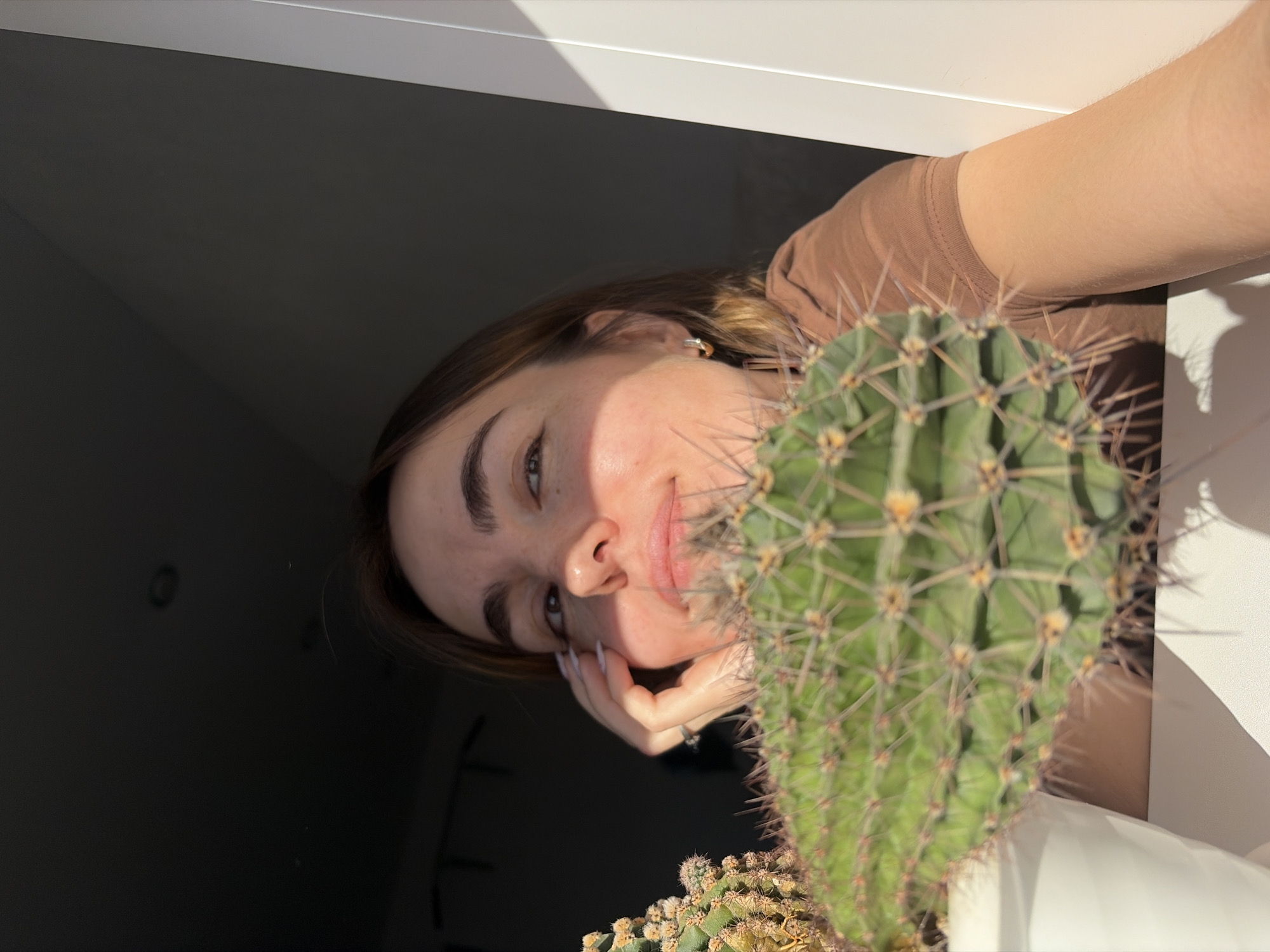 A young woman is posing next to a cactus plant in a white pot. She is wearing a brown shirt and gold earrings. Her right hand is resting on her face, her nails are painted white. She is indoors next to a window, with a dark space visible through the window. Location is unknown, the reference to Sarapul, Russia, is speculative.