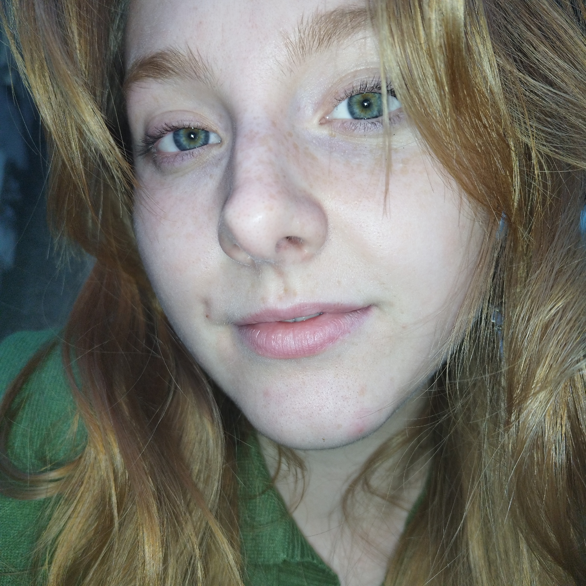 A close-up portrait features a single individual identified as female, positioned slightly off-center, with fair skin and numerous freckles distributed across the nose and cheeks. The subject has long, light reddish-blonde hair that frames the face, with strands partially obscuring the left side. Blue-green eyes are directed towards the viewer, maintaining a direct gaze. The lips are pale pink and slightly parted, displaying a neutral to subtly smiling expression. A minor blemish is visible on the chin. The individual is wearing a dark green garment, consistent with knitted or ribbed fabric, visible at the neckline and shoulders. A small, light-colored earring is partially visible on the subject's right earlobe. The background is uniformly dark and indistinct, providing no discernible environmental details. No specific geographic landmarks or identifying features indicative of a location within Yekaterinburg, Russia, are present in the visual content of the image.