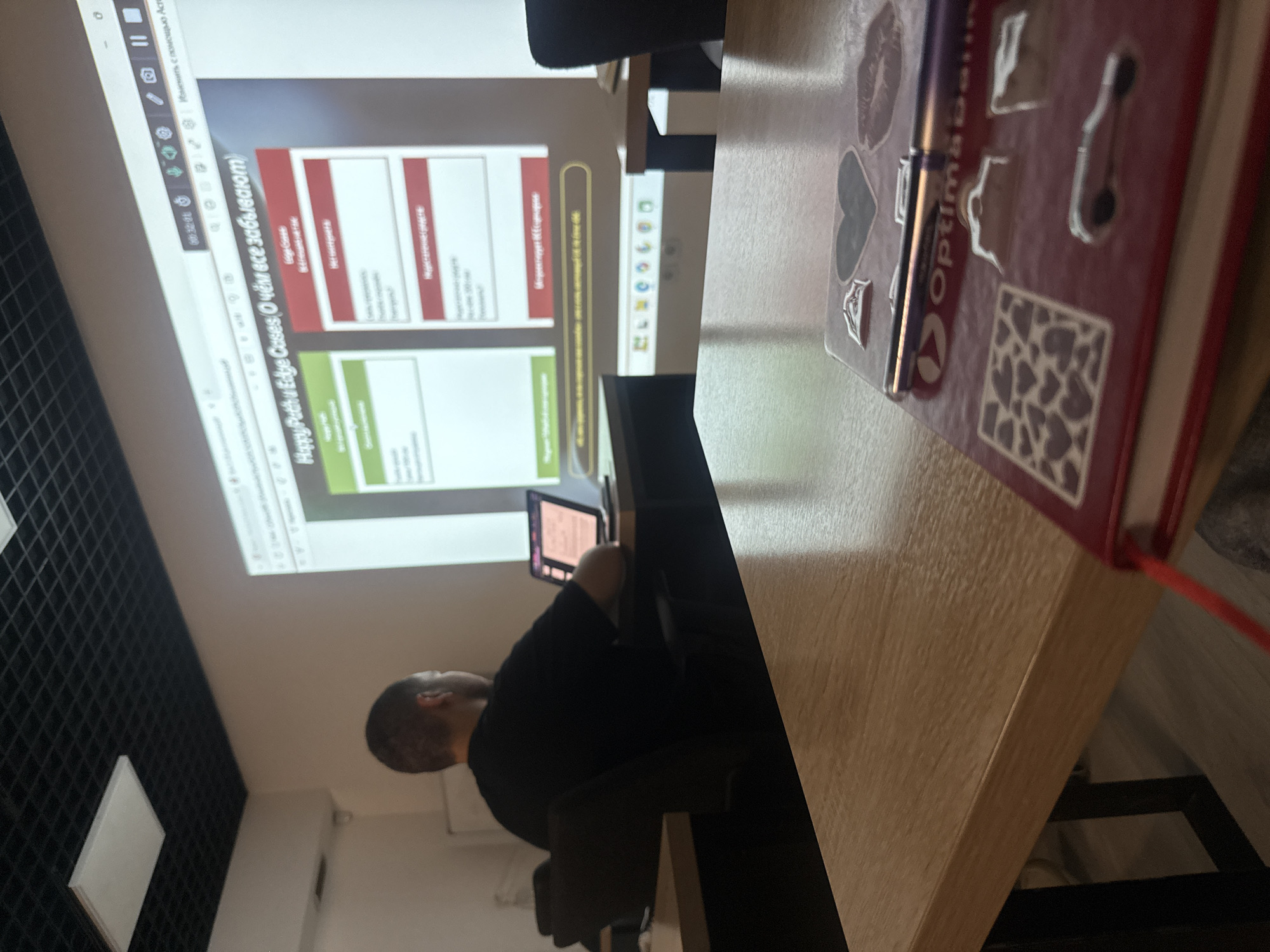 In a dimly lit classroom, a male in a black t-shirt sits at a table facing a projector screen. The screen displays a presentation slide with text in Russian, titled "Happy Path vs. Edge Cases (О чём все забывают)". The man is working on a laptop, its screen illuminated and facing him. The room features a drop ceiling with fluorescent lighting panels. In the foreground, a wooden table is visible with a red notebook and a pen on it, decorated with stickers. The overall atmosphere suggests a lecture or training session. There are no discernible features that place this image in Bishkek City, Kyrgyzstan.