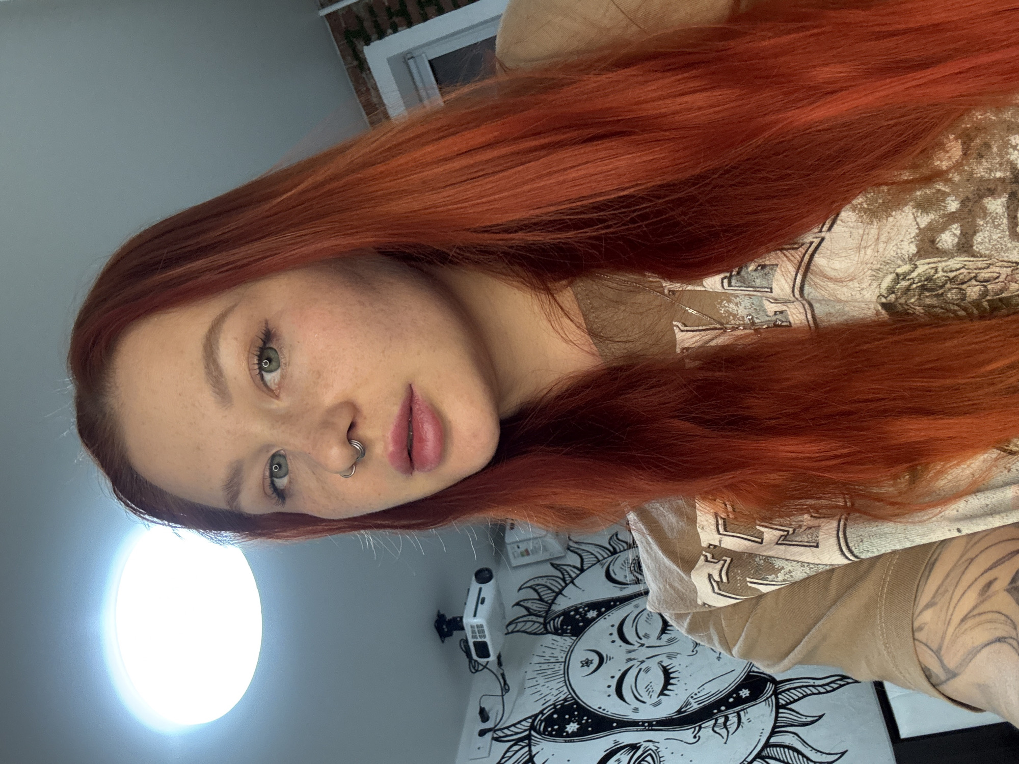 A person with long, red hair and fair skin poses indoors, facing the camera at a slight angle. They have a nose ring and visible freckles. They are wearing a tan-colored shirt with a faded design. Behind the person, a white wall features a black and white tapestry depicting celestial bodies. A light source is also visible, creating a bright spot on the wall. The image does not contain sufficient information to confirm the location as Rostov-on-Don, Russia.