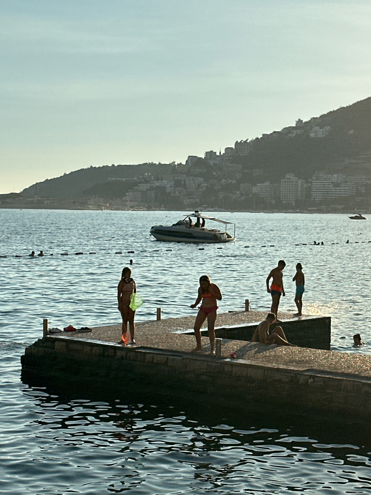 An elevated, silhouetted view of a concrete pier extending into the Adriatic Sea is captured at dusk. Several individuals, including children, are present on the pier, some in swimwear. One child stands with a green net, appearing to prepare for fishing. Another child sits on the edge of the pier. A boat is visible in the background, cruising on the water. The coastline behind the sea is lined with buildings, suggesting an urban setting. The scene takes place in the city of Budva, Montenegro, as evidenced by the distinct mountainous backdrop and urban development characteristic of the area. The lighting suggests late afternoon or early evening, with the sun casting long shadows.