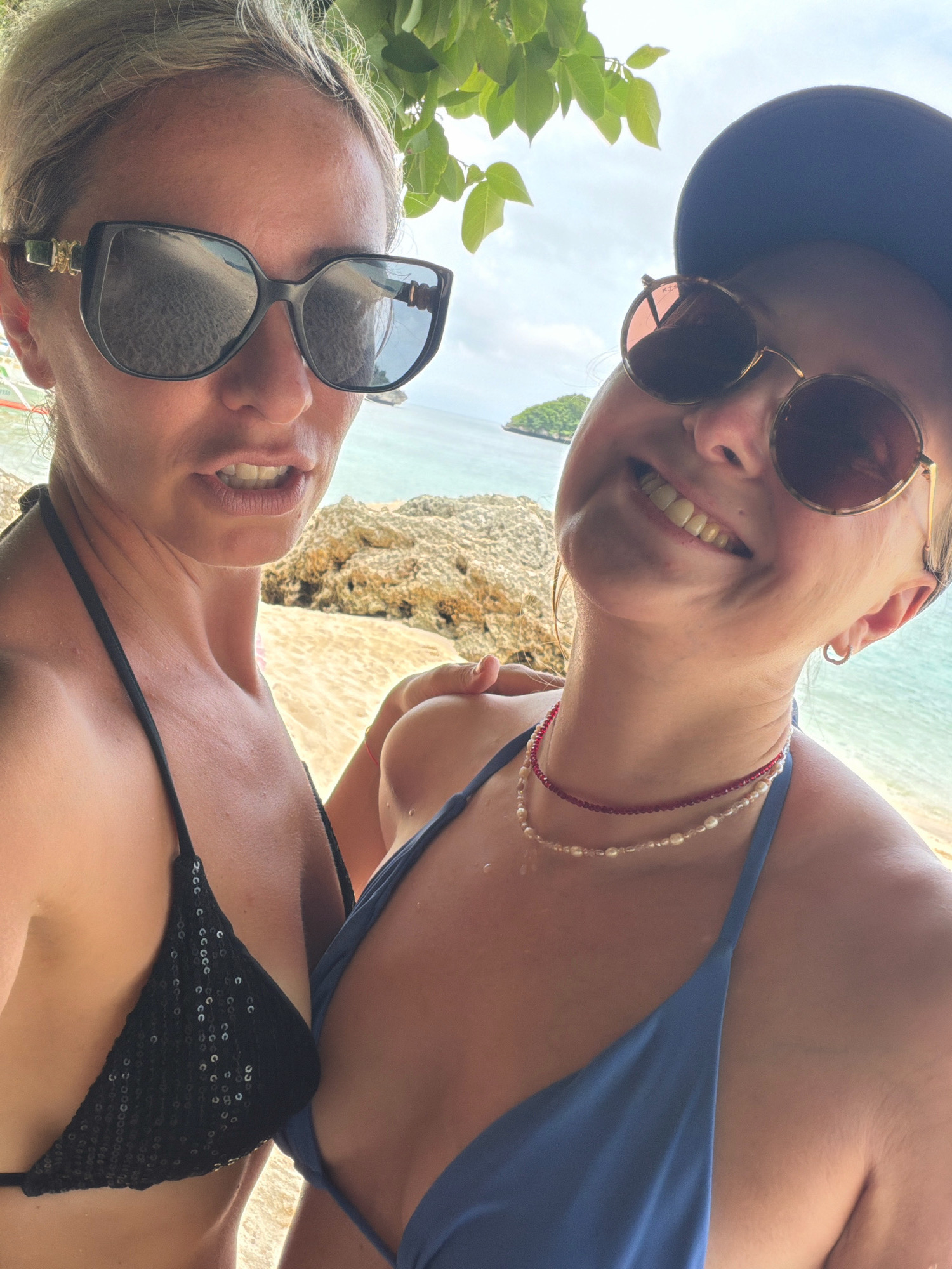 Two women are taking a selfie on a beach. The woman on the left is wearing large black sunglasses, a black sequined bikini top, and has blonde hair pulled back. She is looking directly at the camera with her mouth slightly open. The woman on the right is wearing round sunglasses, a blue bikini top, and a dark baseball cap. She is smiling and looking towards the camera. Both women appear to be on a sandy beach with rocks and the ocean in the background. The location is likely Boracay Island in Malay, Philippines, given the sandy beaches, clear blue water, and tropical foliage visible in the background, characteristic of the island's many beach areas.