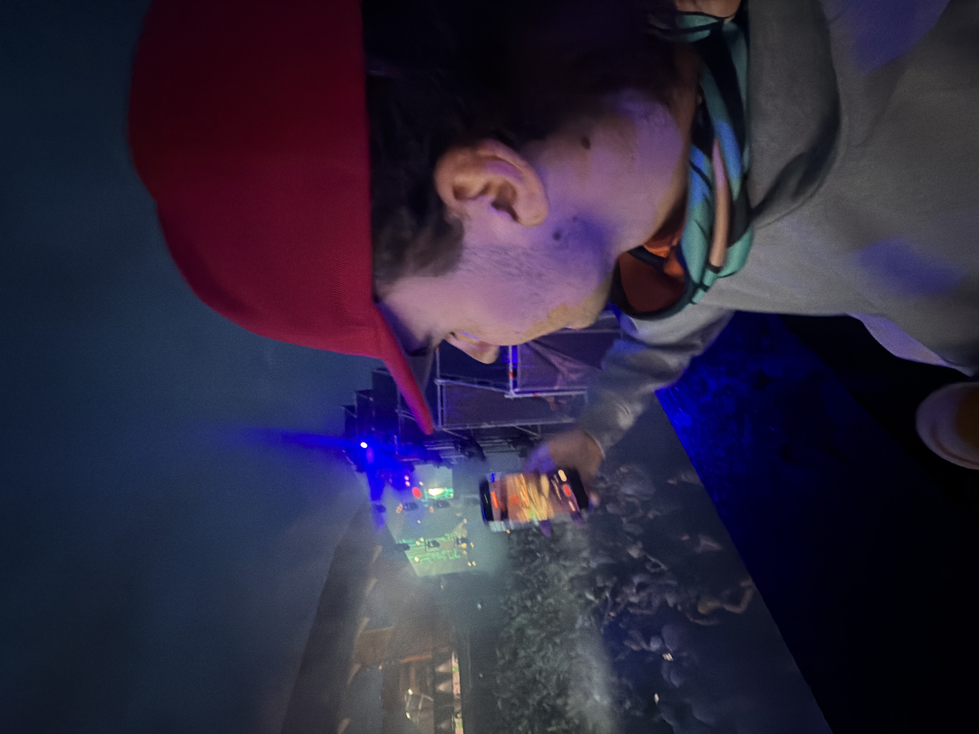 An elevated, candid shot captures a male individual from the right side at a large-scale nighttime music event in Sant Adrià de Besòs, Spain. The subject wears a bright red baseball cap reversed, a grey hooded sweatshirt, and a multicolored neck gaiter displaying teal, orange, and blue geometric patterns. Light stubble and two distinct dark moles are visible on his right cheek. He holds a black smartphone in his extended right hand, with the illuminated screen displaying a live feed or image of the event. His gaze is directed towards the activity below.
In the mid-ground, a large stage structure is prominent, featuring multiple LED screens displaying abstract green and red visuals. Intense blue and green stage lights project beams through atmospheric fog or smoke, illuminating a dense crowd of attendees. To the far left, dimly lit architectural structures, possibly part of a stadium or venue, are discernible. A metallic truss system is visible to the right of the stage. The sky above is dark blue, indicating a night setting. The scene is characterized by dynamic artificial lighting and a large, engaged crowd.