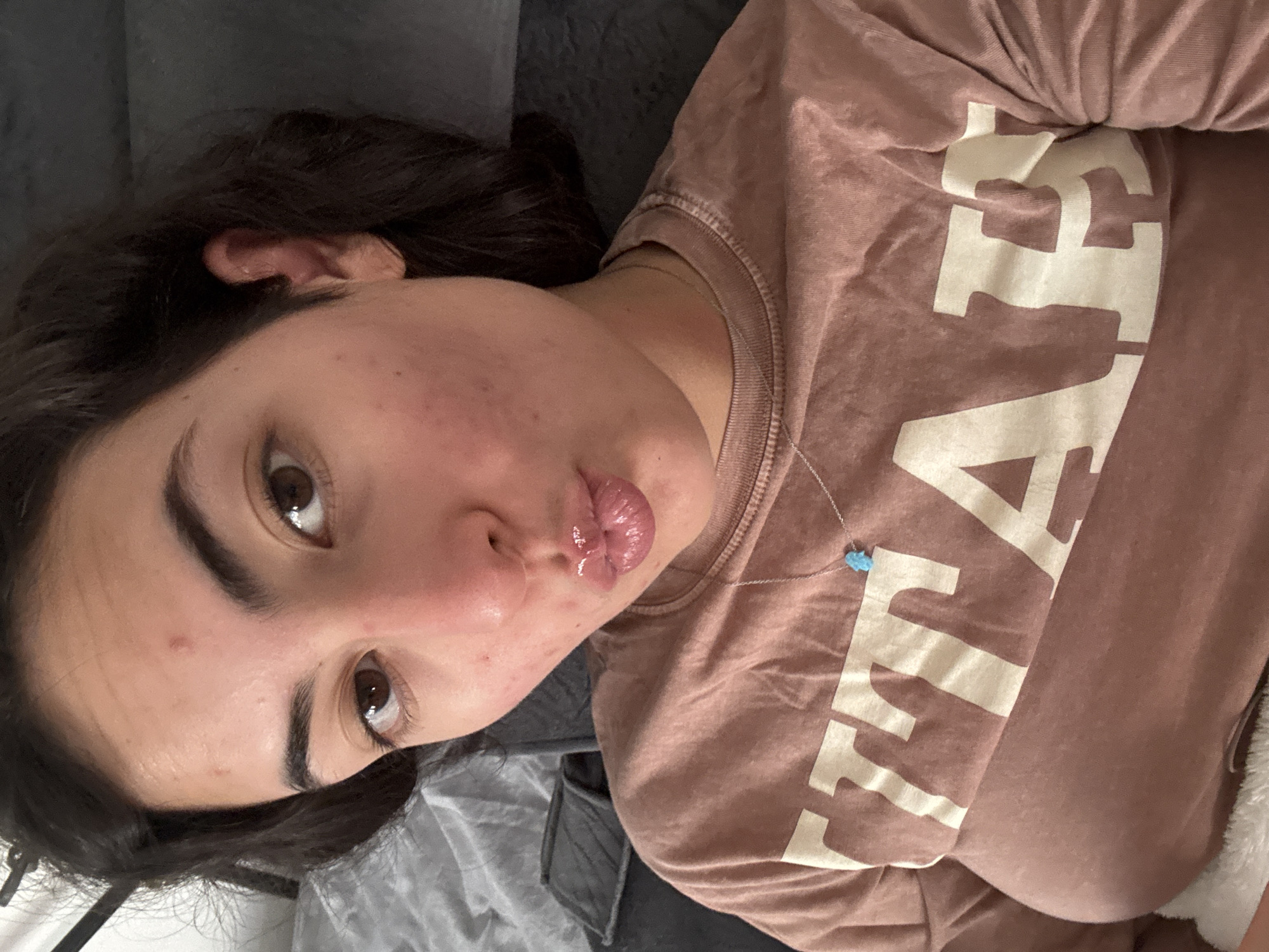 A young woman is lying on a gray blanket or sheet, wearing a mauve-colored shirt with large white letters on it. She has dark brown hair and is looking at the camera with a slight pout. The scene is indoors and the lighting appears to be ambient. A delicate necklace with a blue charm is visible around her neck. Her skin shows some blemishes. Without additional information, no specific location in Puebla, Mexico, can be determined.