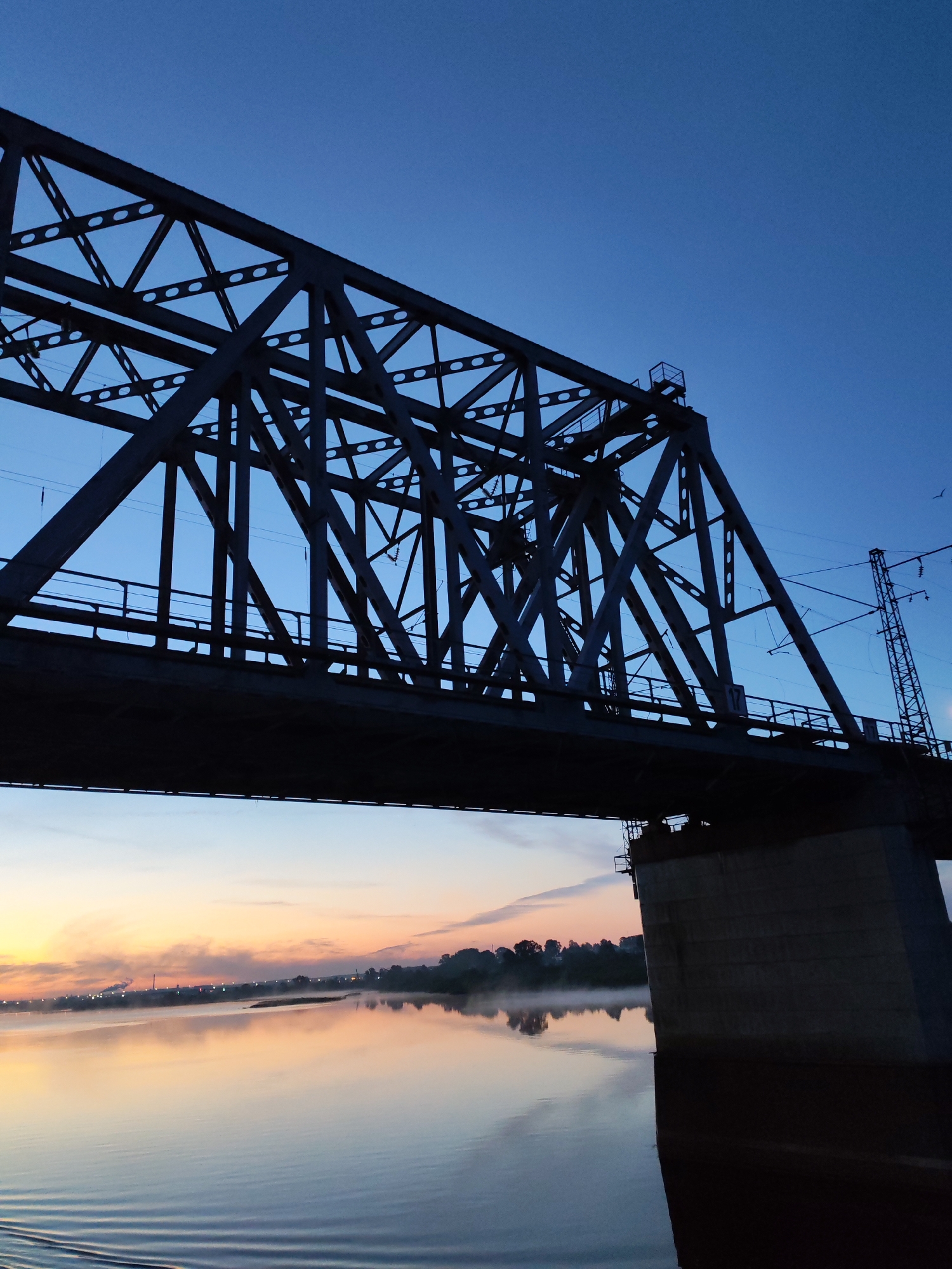 The image depicts a steel truss bridge, silhouetted against a gradient sky transitioning from orange near the horizon to deep blue above. The bridge supports a railway track and has a concrete support structure visible on the right. Below the bridge, a calm body of water reflects the colors of the sky and the faint lights of the distant shoreline, which is obscured by a band of mist. The location is identified as Tarkanovo, Russia.