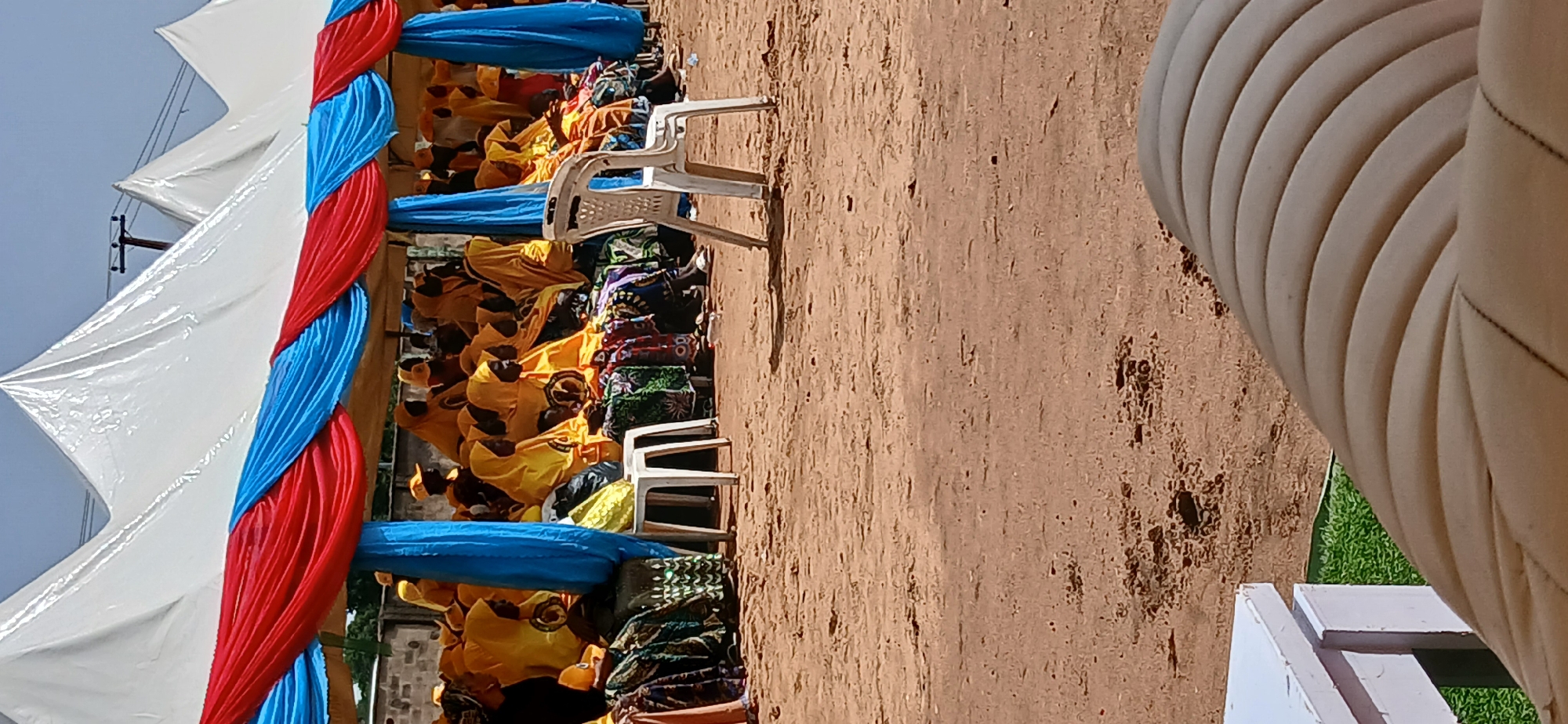 In Rigachikun, Nigeria, under a white tent adorned with red and blue fabric, a group of people, predominantly women, are seated. They are dressed in yellow attire and headscarves, with some wearing matching yellow caps. The ground beneath them is sandy. In the foreground, a white plastic chair stands empty. To the left, a white table is partially visible, with a patch of green grass behind it. The scene suggests an outdoor gathering or event.