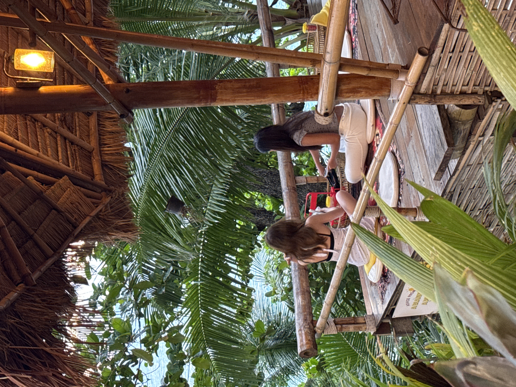 In Karon, Thailand, two young women sit on a wooden platform under a thatched roof, surrounded by lush green palm trees and overlooking the turquoise ocean. The platform, constructed from bamboo and wood, is raised and appears to be part of an outdoor dining area or seating space. Both women are dressed casually. The woman on the left wears a white dress with black polka dots and a black bikini top. She is facing away from the camera, looking towards the sea. The woman on the right, also facing away from the camera, wears a patterned grey top and white pants. She is looking down at something in her hands. On the platform, there are cushions, woven mats, and a basket containing food items. A warm light bulb hangs from a wooden beam, casting a soft glow. The overall scene depicts a relaxed, tropical setting with a view of the natural beauty of Karon.