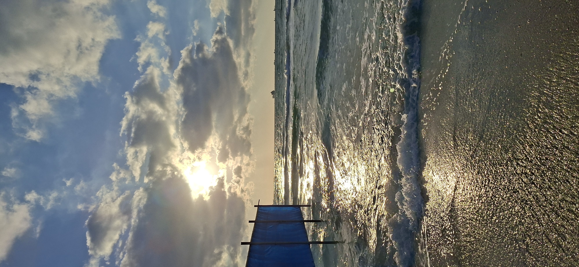 The image depicts a serene seascape with a bright sun shining through clouds over the ocean. In the foreground, a blue barrier, possibly a windbreak or enclosure, stands on the wet sand at the edge of the water. The sun's rays create a shimmering reflection on the surface of the gentle waves as they approach the shore. Several ships are visible on the horizon, suggesting maritime activity in the distance. The sky is a mix of blue and grey clouds, with the sun partially obscured, casting a warm, diffused light over the scene. The beach appears empty of people, contributing to the tranquil atmosphere.