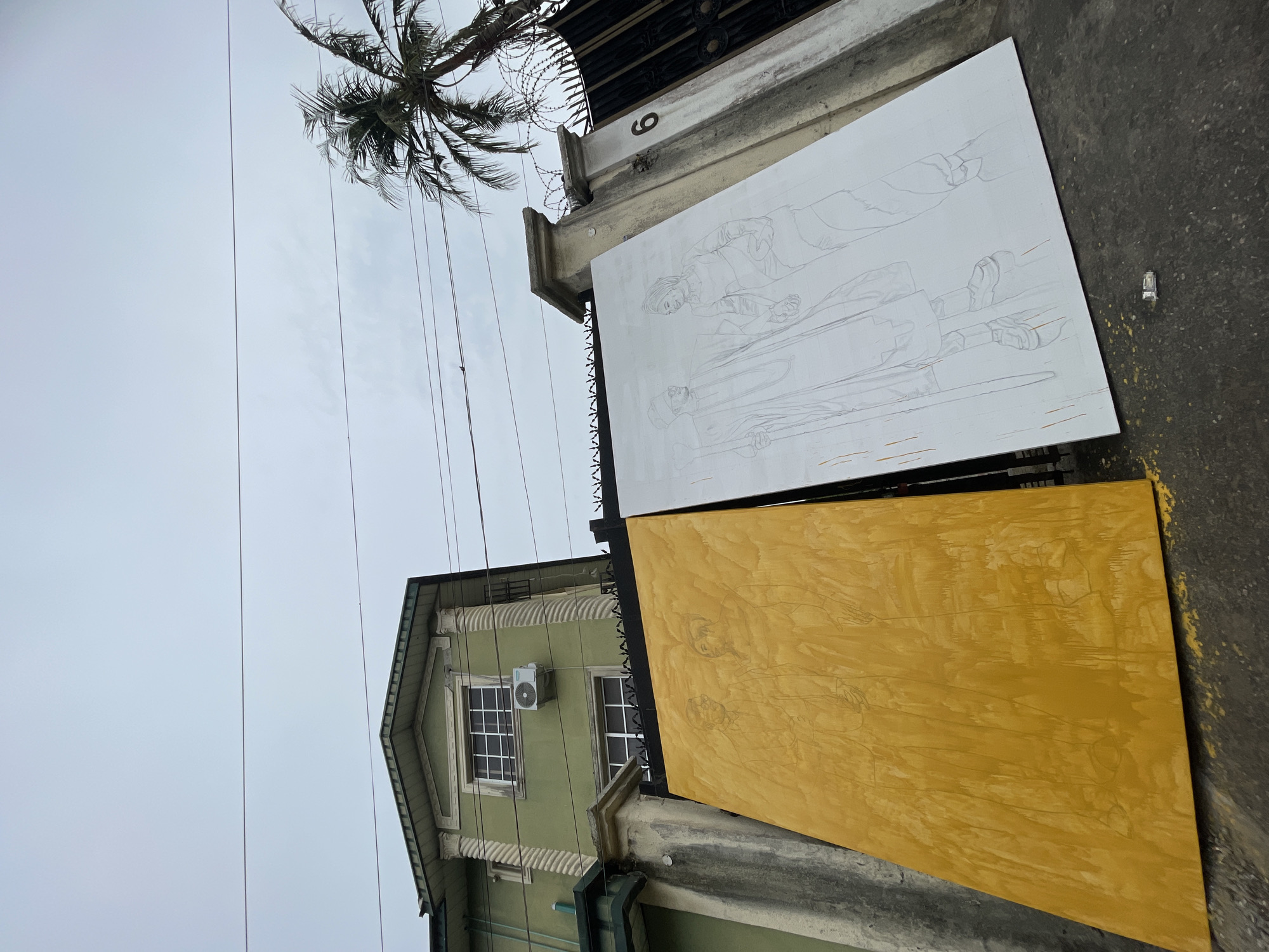 The image captures an outdoor scene in Ado, Nigeria. Two large rectangular boards, possibly canvases or panels, lean against a wall or structure. The upper board is white with a sketch of figures on it. The lower board is yellow, also bearing a sketched image. Above the boards, a wall is visible with the number "6" affixed to it. A palm tree extends from behind the wall, with overhead electrical wires stretching across the sky. In the background, there is a two-story building painted in a green hue with white-framed windows and an external air conditioning unit. The sky is overcast.