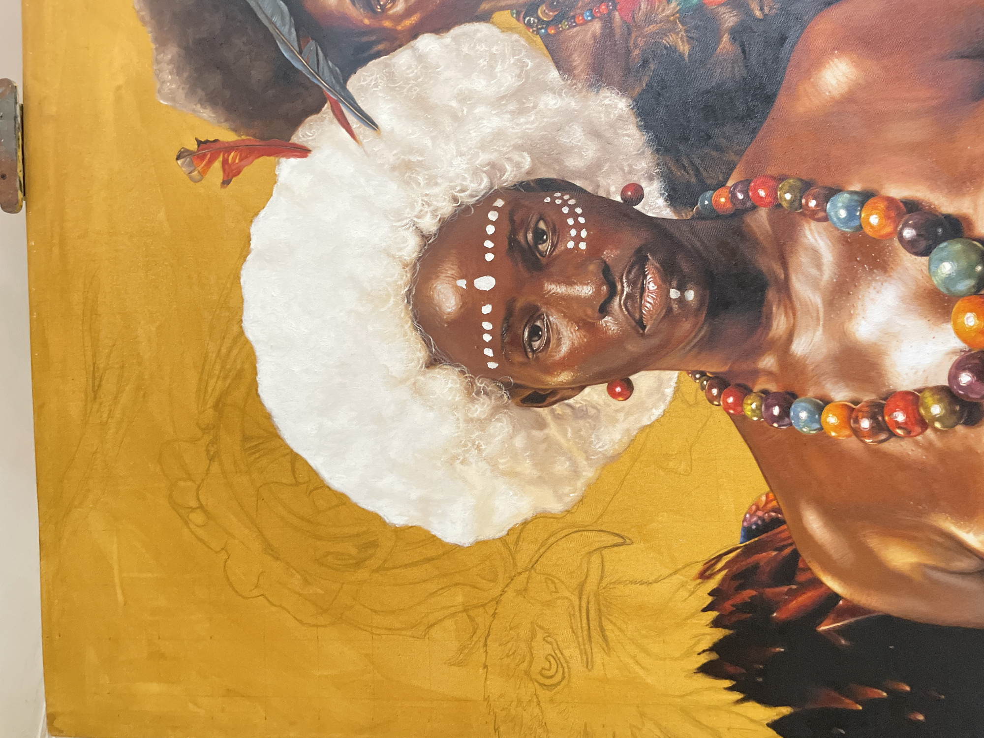 The image shows a partial painting of a woman with dark skin, wearing white dots on her face and a large, white, ruffled collar. She has a necklace of colorful, round beads. The background is a golden yellow, with a sketched outline of another figure. Part of another person's head is visible in the upper portion of the painting. The location is Ado, Nigeria.