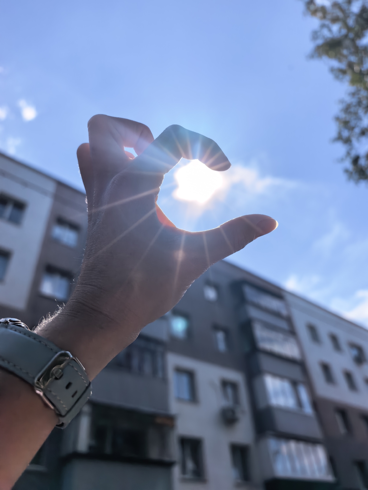 A hand is held up in front of the sun, creating a crescent shape. The sun is bright and flares in the image. The hand is wearing a light blue watch. In the background, there is a blurred multi-story building with windows and balconies. The sky is blue with some clouds. A few branches of a tree are visible in the upper right corner. The image appears to have been taken outdoors during the day. The location is not specified in the image.