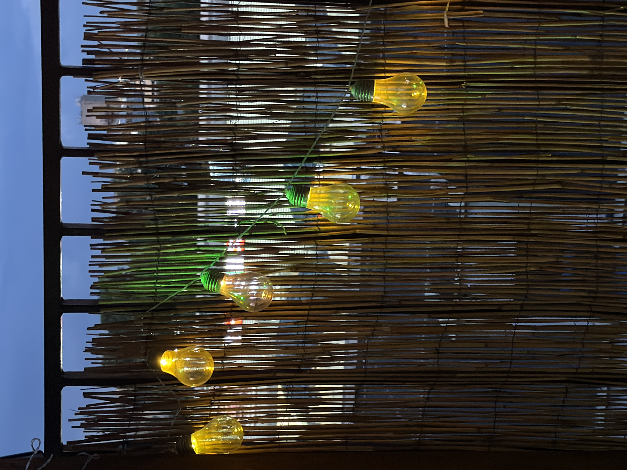 A string of at least eight yellow-orange LED bulbs, shaped like traditional incandescent bulbs, is illuminated and strung diagonally downwards from left to right in front of a bamboo screen. Some of the bulbs feature a distinct green base. The screen, serving as a privacy barrier, is constructed from numerous vertical brown and tan bamboo or reed sticks, held together by thin horizontal ties. Through the narrow vertical gaps in the bamboo screen, the blurred lights of an urban or suburban environment are visible in the background, appearing as streaks of white, red, and orange. In the upper midground, partially obscured by the screen and a dark metal railing, a light-colored, flat-roofed building or structure is discernible against a dark blue twilight sky. The viewpoint suggests an elevated outdoor position, such as a balcony or patio, in the city of Unknown, Unknown. No individuals are present.