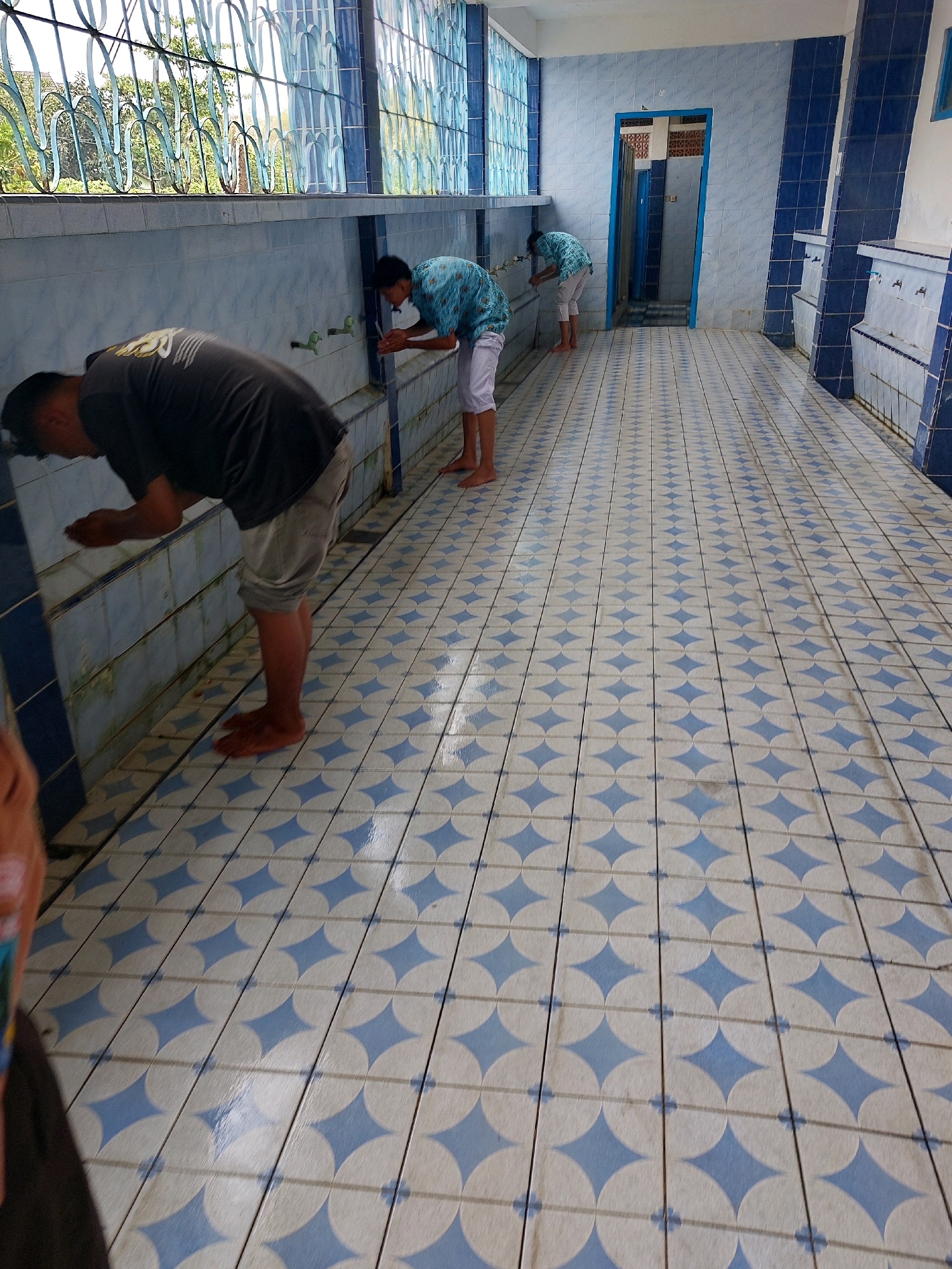 This image captures a scene in what appears to be a public ablution area, possibly at a mosque or a religious school in Sumberrejo, Indonesia. Three young men are visible, each engaged in washing their hands and faces at a row of faucets. The floor is covered with distinctive blue and white patterned tiles, with some areas appearing wet, indicating recent use. The walls are tiled in a lighter blue, and a decorative metal grate with a wavy design runs along the top portion of the wall, allowing natural light to enter from outside. In the background, a doorway with a blue frame leads into another room. The overall atmosphere suggests a functional space dedicated to ritual purification.
