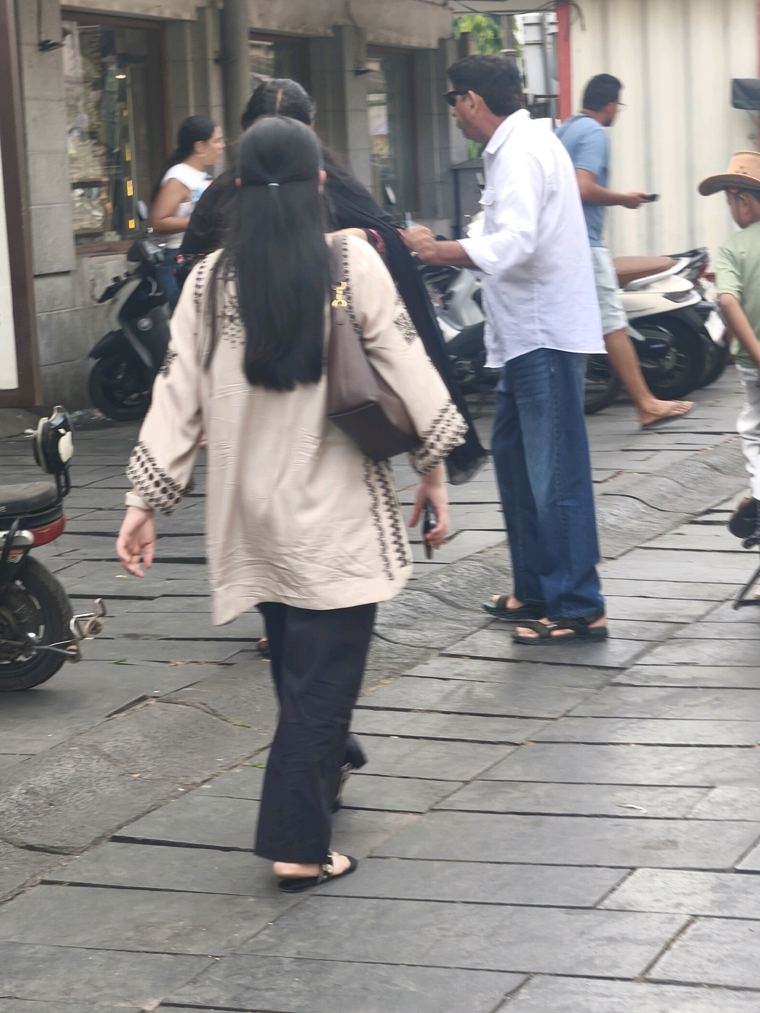 A group of people are visible on a pedestrian walkway paved with large, dark gray rectangular stones. In the foreground, a woman with long black hair walks away from the camera, wearing a light beige tunic with decorative black embroidery on the sleeves and hem. She carries a brown handbag over her right shoulder. To her left, a black motorcycle is parked. Behind her, a man in a white long-sleeved shirt and blue jeans walks towards the right, looking to his left. Another man in a blue t-shirt and light shorts walks away from the camera on the right side of the scene, holding a mobile phone. Further to the right, a person wearing a light green t-shirt and a brown hat is partially visible. The environment appears to be an urban setting with a building in the background. The overall scene suggests a busy street or public area in West Jakarta, Indonesia.