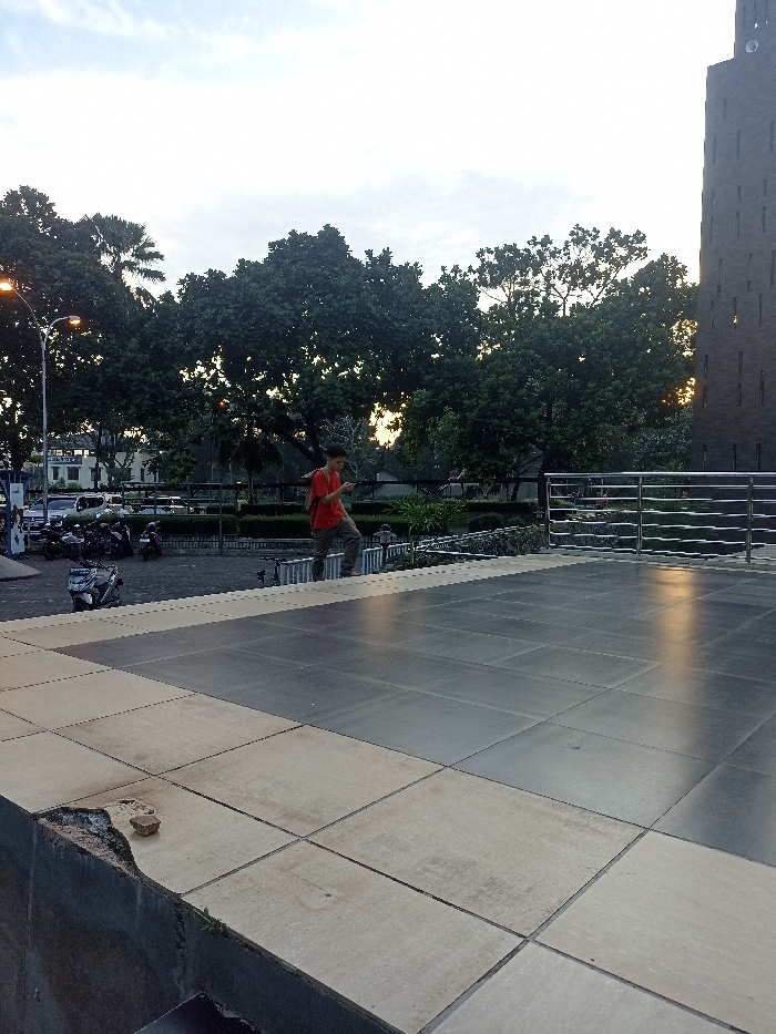 The image shows a man in a red shirt walking toward a building with a large black paved area in front. The man is holding a smartphone in his left hand. The building is made of brick and has a flat roof. There are trees and bushes in the foreground and background. There are parked motorcycles to the left of the image. There is a fence made of metal bars to the right. The image is taken from the perspective of someone standing on the paved area. The coordinates provided (latitude: -6.931, longitude: 107.776) indicate that the image was taken in Jatinangor, Indonesia. The sky is overcast, but the lighting is bright.