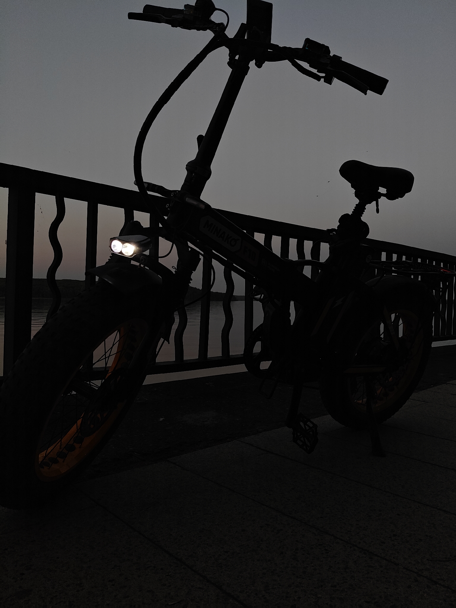 The image depicts a Minako F18 electric bicycle parked on a paved surface, likely a sidewalk or pathway. The bike is silhouetted against a dimly lit background, suggesting either twilight or a low-light condition. The bike is leaning against a dark railing, with what appears to be a body of water visible in the background through the railing. The bicycle's headlight is illuminated, providing the only direct source of light in the image. The tires are wide and partially visible, revealing an orange rim. The location is inferred to be the city of Chaikovsky, Russia, based on the bike model "Minako F18" potentially being popular in the region. No people are visible in the image.