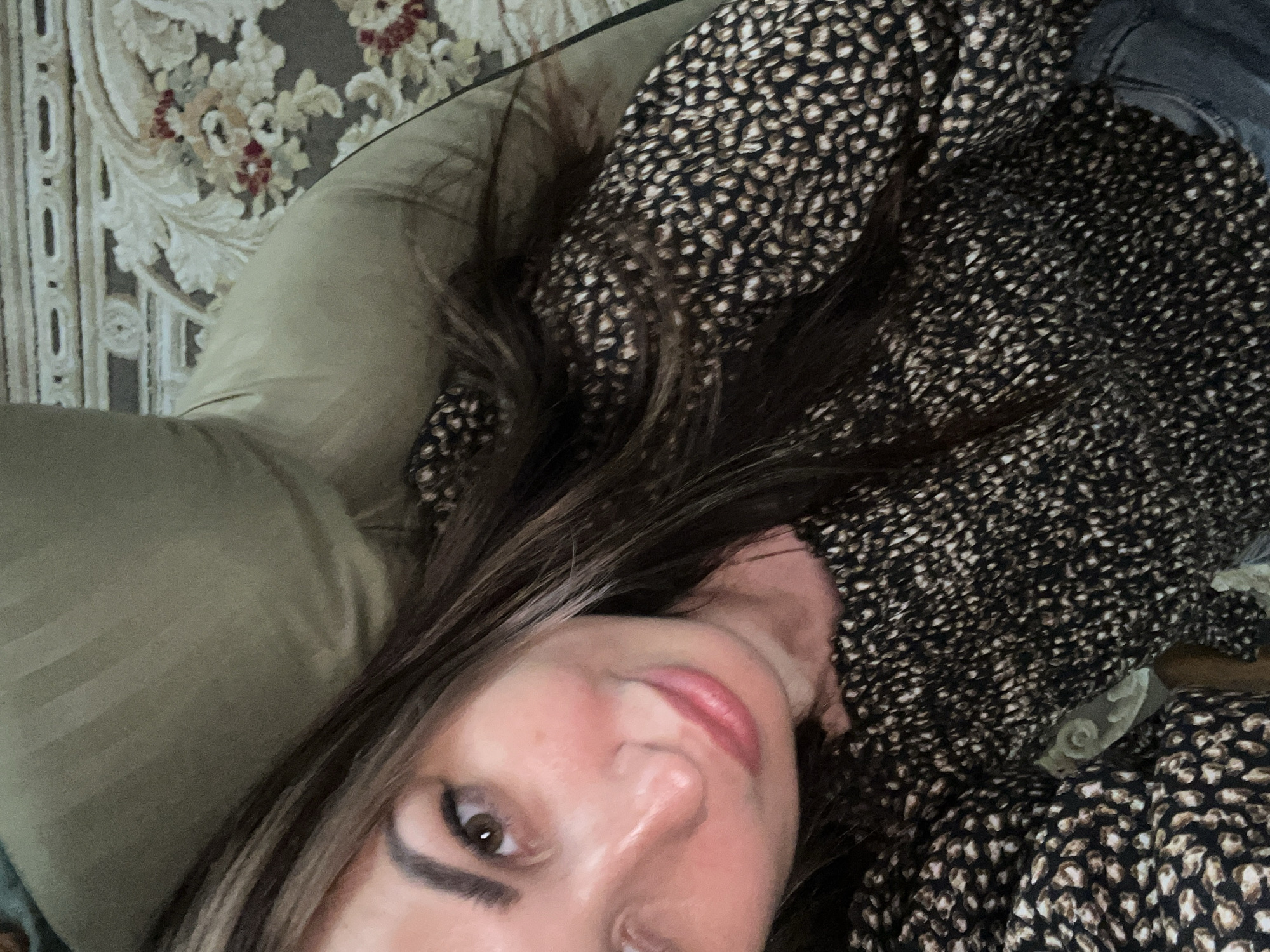A woman is lying on a couch, looking up at the camera. She has long, dark hair with some lighter streaks and is wearing a dark top with a small, repeating pattern. Her face is tilted, and she has makeup on, including pink lipstick. The background shows part of the couch and a rug with a floral pattern. The lighting is dim.