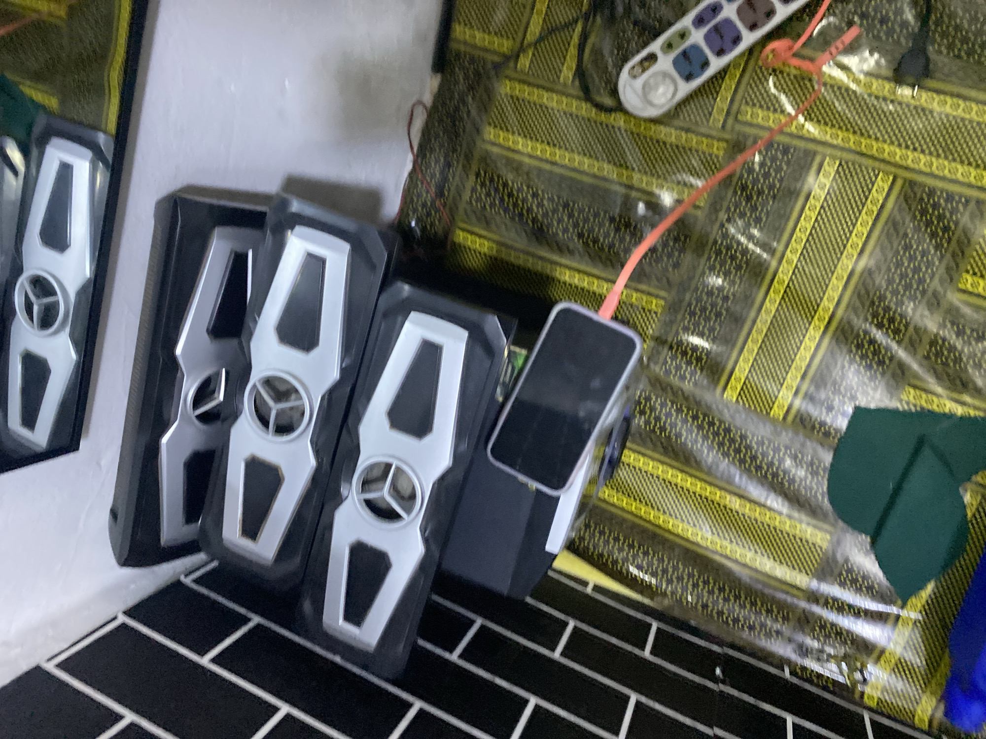 The image depicts a collection of black and silver objects stacked vertically, possibly speakers or electronic devices, placed on a surface covered with a yellow and black patterned material. A smartphone, connected to a red cable, rests on top of one of the stacked objects. To the right, a white remote control with several buttons is visible. In the background, a wall with black and white tiled sections can be seen. The overall scene appears to be indoors, possibly in a residential setting in Jalingo, Nigeria, given the typical floor coverings and proximity to a tiled wall often seen in such environments. The lighting suggests an indoor setting with artificial light sources.
