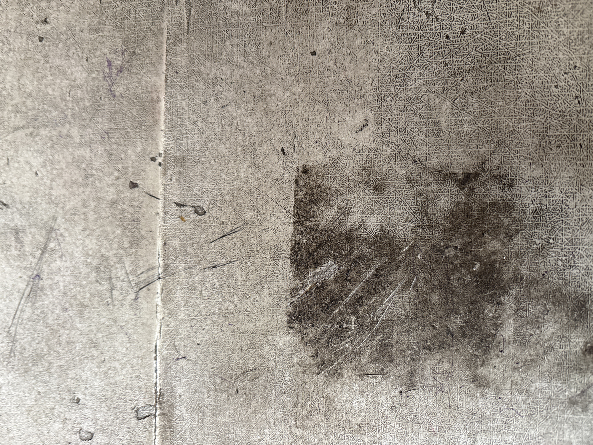This is a close-up, overhead shot of a textured, grey surface that appears to be worn and dirty. The surface has a fine, repeating pattern embossed into it, resembling a grid or a woven texture. There are visible scratches and scuff marks across the surface, with a prominent dark stain in the lower left quadrant of the image. Small dark specks, possibly dirt or debris, are scattered throughout. A thin horizontal line, suggesting a seam or a crease, runs across the middle of the image, dividing the surface into two slightly different textured sections. No people, actions, or specific objects are discernible beyond the texture and imperfections of the surface itself. The location in Palimanan, Indonesia, is not evident from the visual information provided in the image.