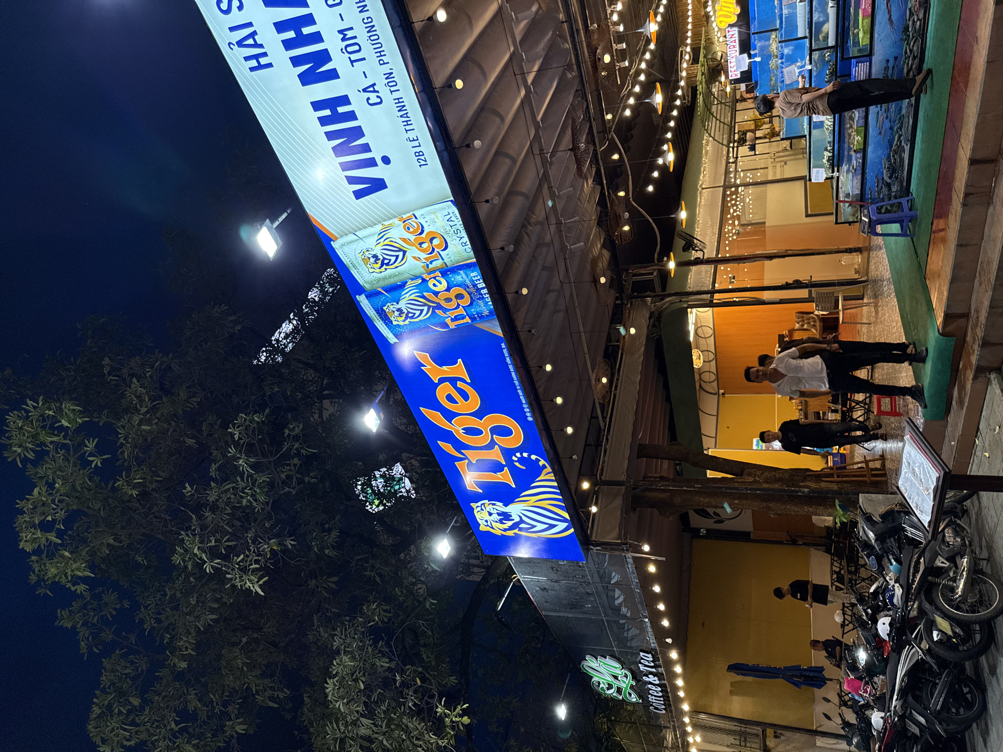 The image depicts a nighttime scene on a street in Nha Trang, Vietnam. Dominating the scene is a large, illuminated blue sign for "Tiger Beer" featuring a tiger graphic and two cans of the beverage. Adjacent to it, a long white sign reads "VINH NHA" in large blue letters, with smaller text below indicating "CÁ - TÔM - GHẸ" and an address, "12B LÊ THÁNH TÔN, PHƯỜNG PHƯỚC, THÀNH PHỐ NHA TRANG, KHÁNH HÒA".
Below these signs, a covered outdoor dining area is visible, illuminated by string lights and pendant lamps. Several patrons are seated at tables within this area. In the foreground, motorcycles are parked along the sidewalk. Further to the right, a seafood restaurant is evident with tanks displaying live seafood. People are interacting with the seafood tanks and the general environment appears to be a lively commercial street at night. The presence of trees suggests an urban setting with some greenery.