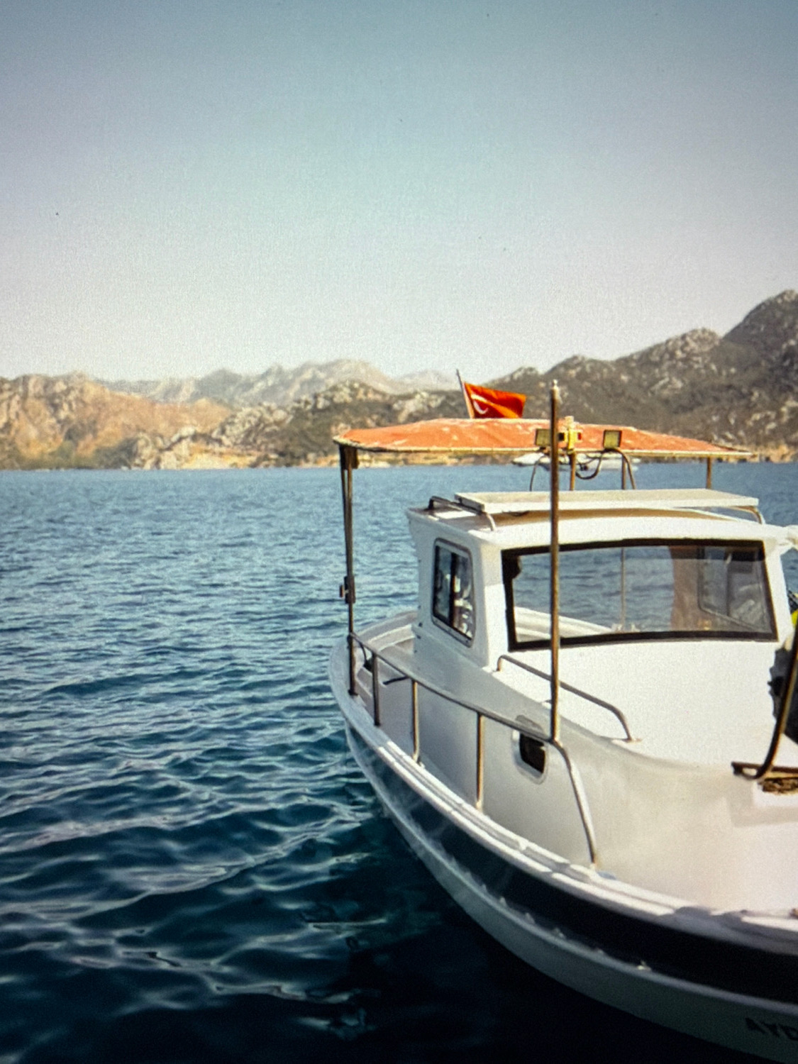 A white boat with a red awning is docked on a body of water. The boat has a cabin with windows and railings. A Turkish flag is visible on the awning. In the background, there are mountains and a clear sky. The image does not provide any information or context that would allow for identification of the location in the city of Kafr Yasif, Israel.